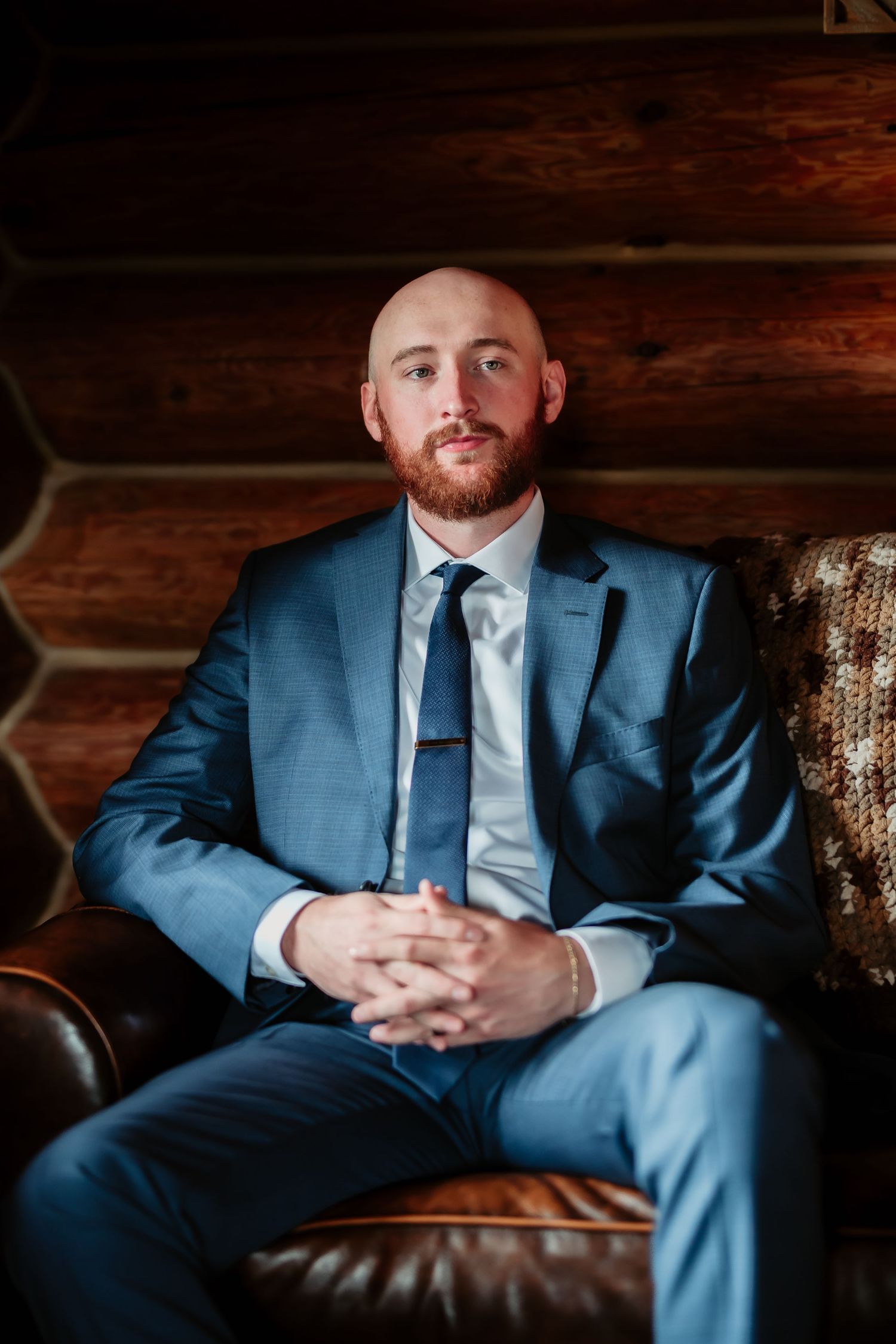 A person in a fitted blue suit and tie sits on a brown leather couch inside a log cabin.