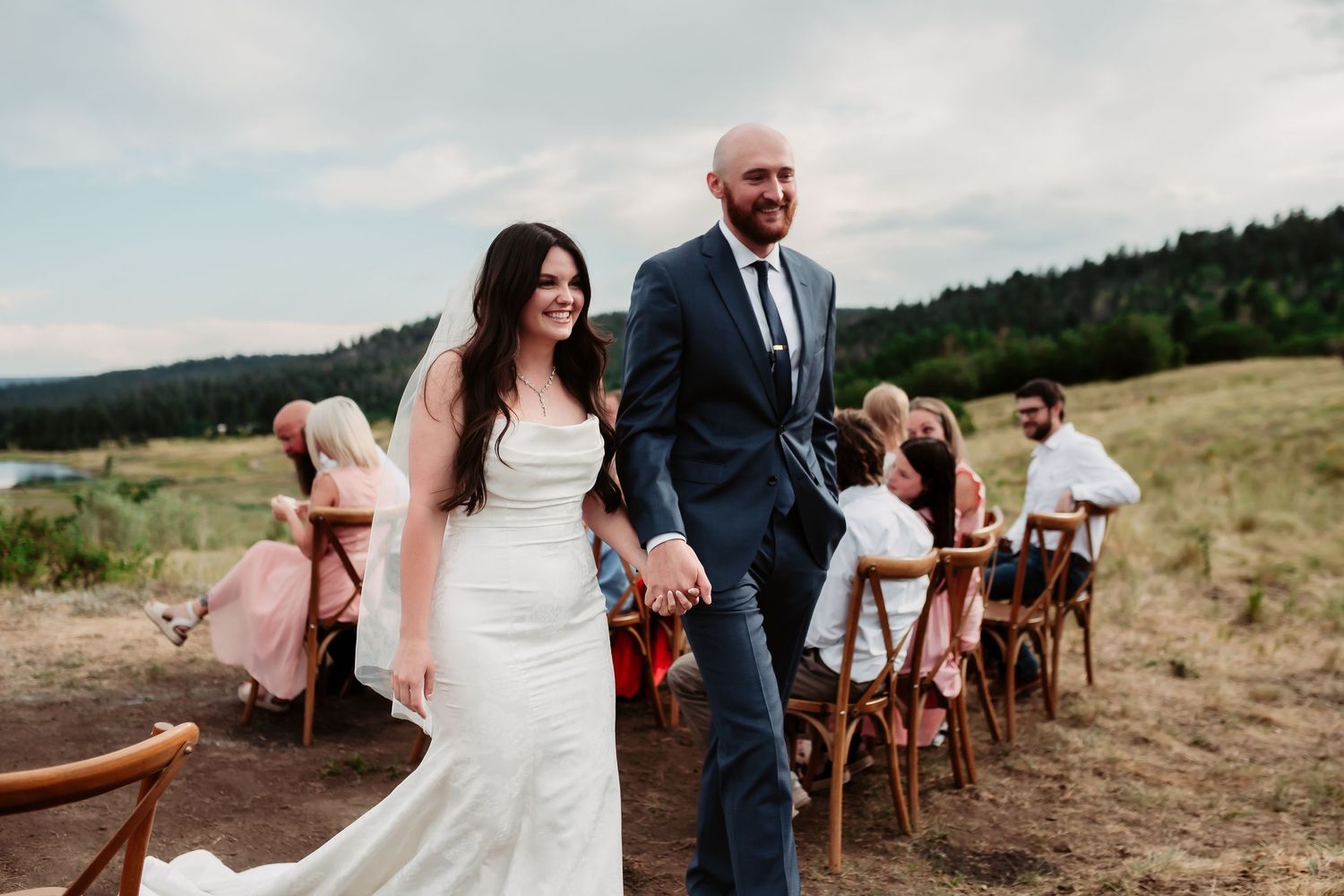 A newlywed couple walks down the aisle at their outdoor mountain wedding while guests look on.