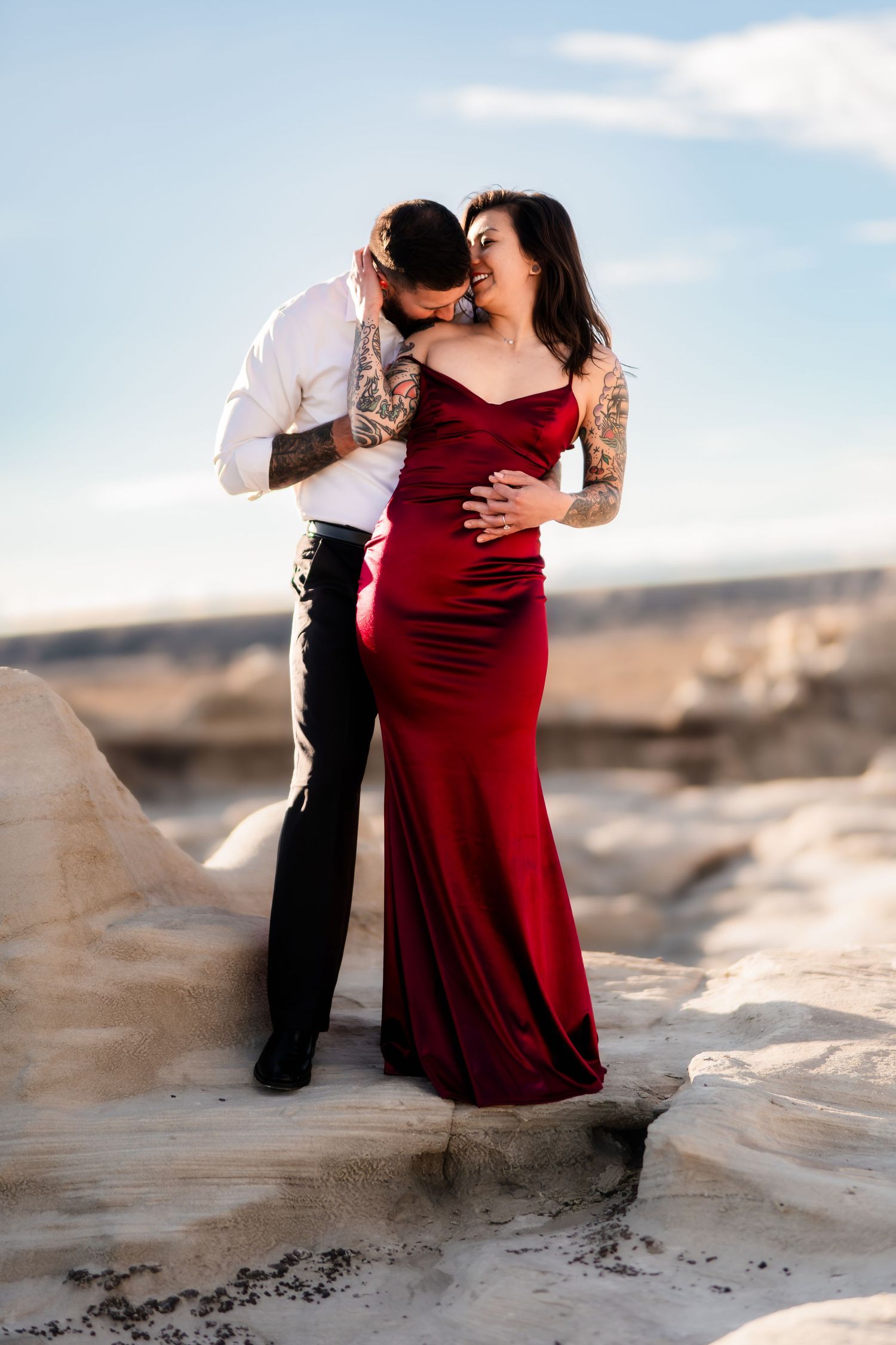 A romantic couple poses on rocky terrain, with a woman in a red velvet gown and a man in formal attire embracing.