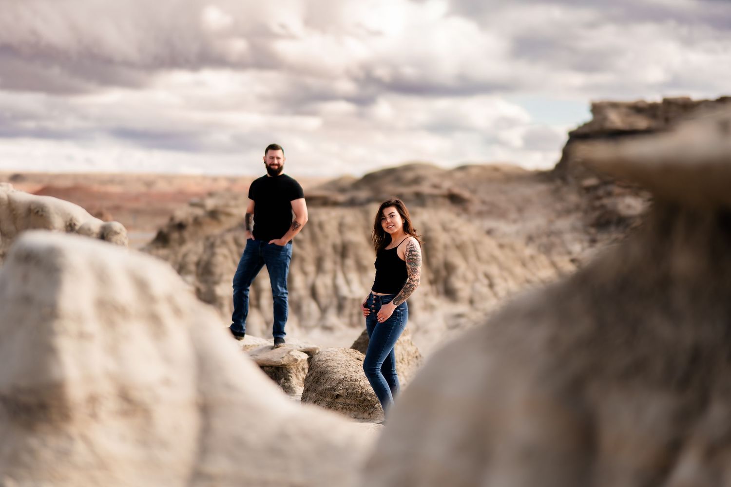 Two people stand on dramatic rock formations in a desert badlands landscape under moody cloudy skies.
