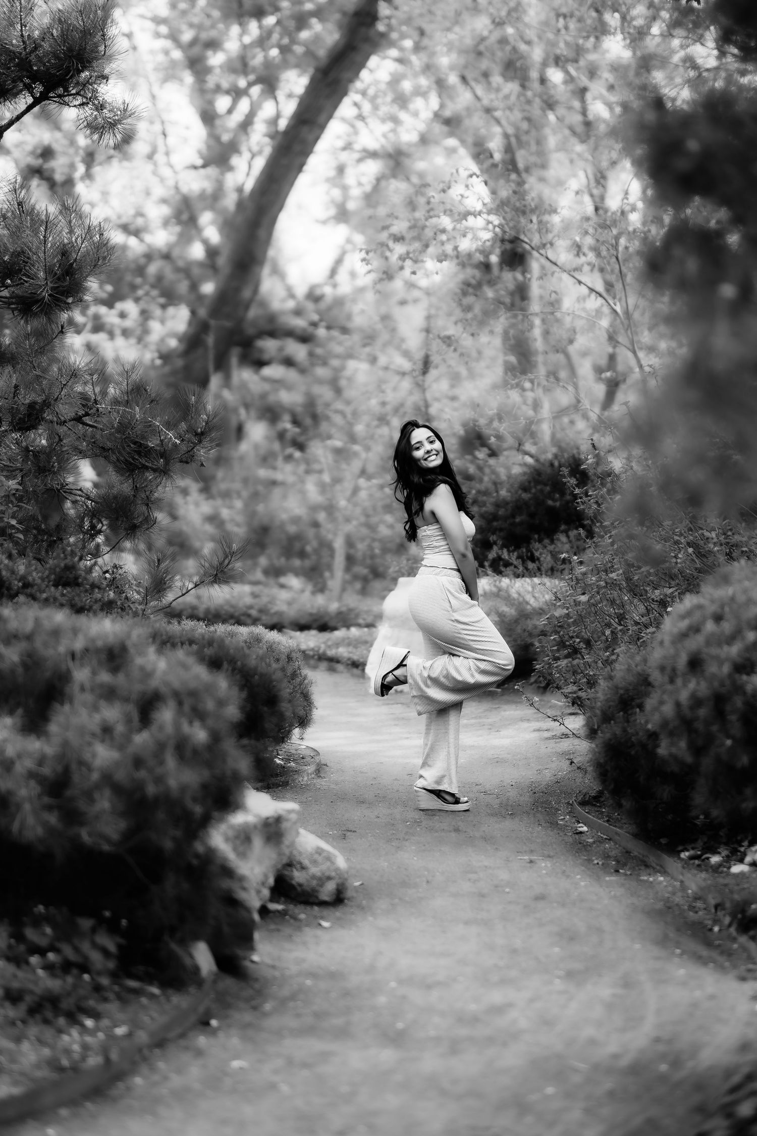A black and white photo of someone in flowing clothes dancing on a garden path surrounded by trees.