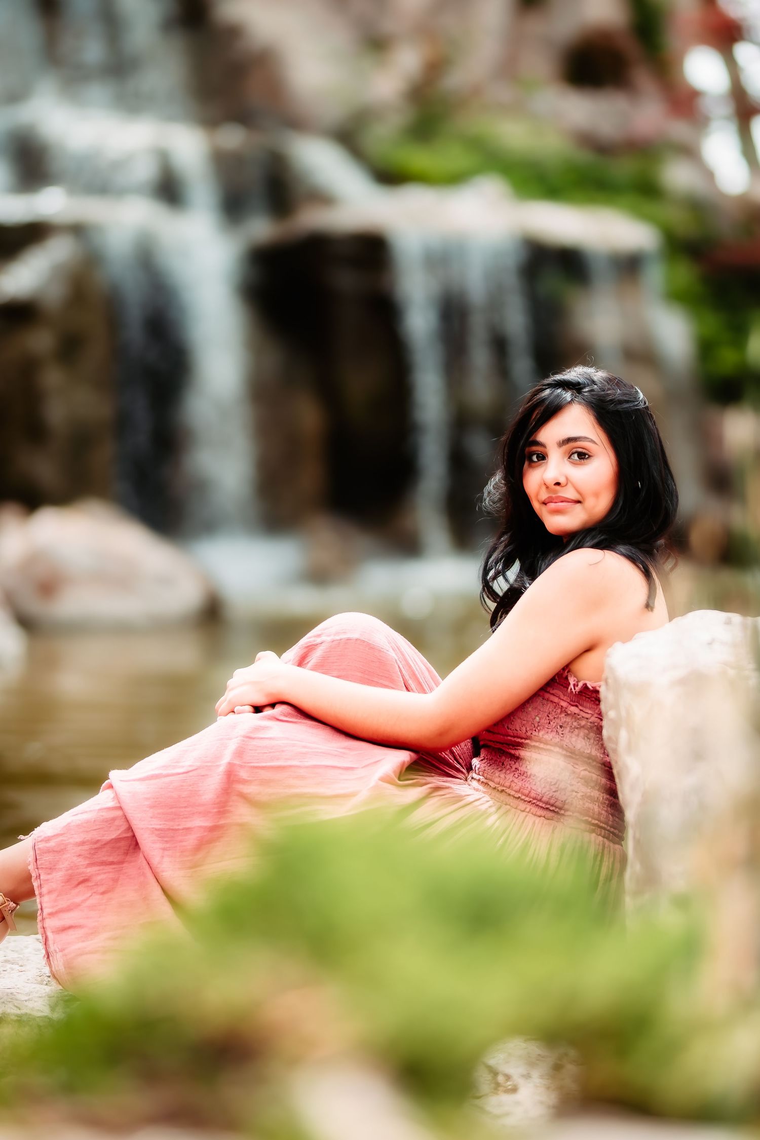 Woman in pink dress poses by cascading waterfall in garden setting.