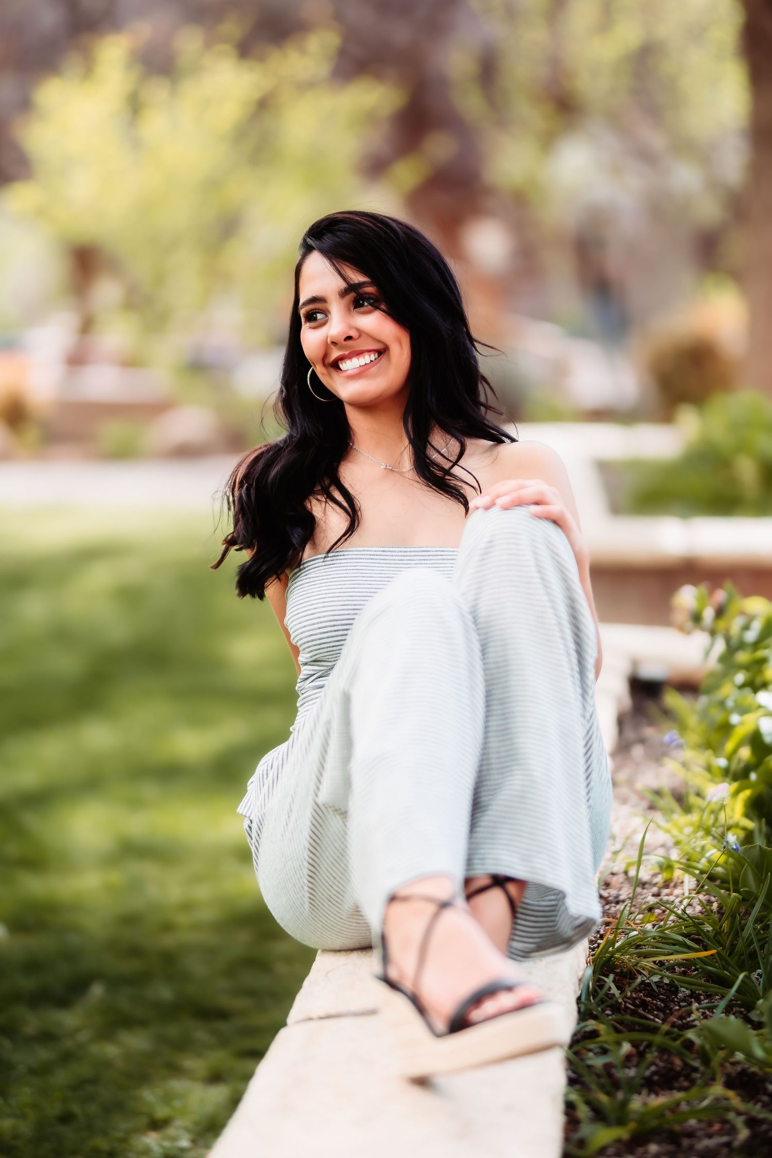 Person in casual attire poses outdoors on a garden path surrounded by lush greenery on a sunny day.