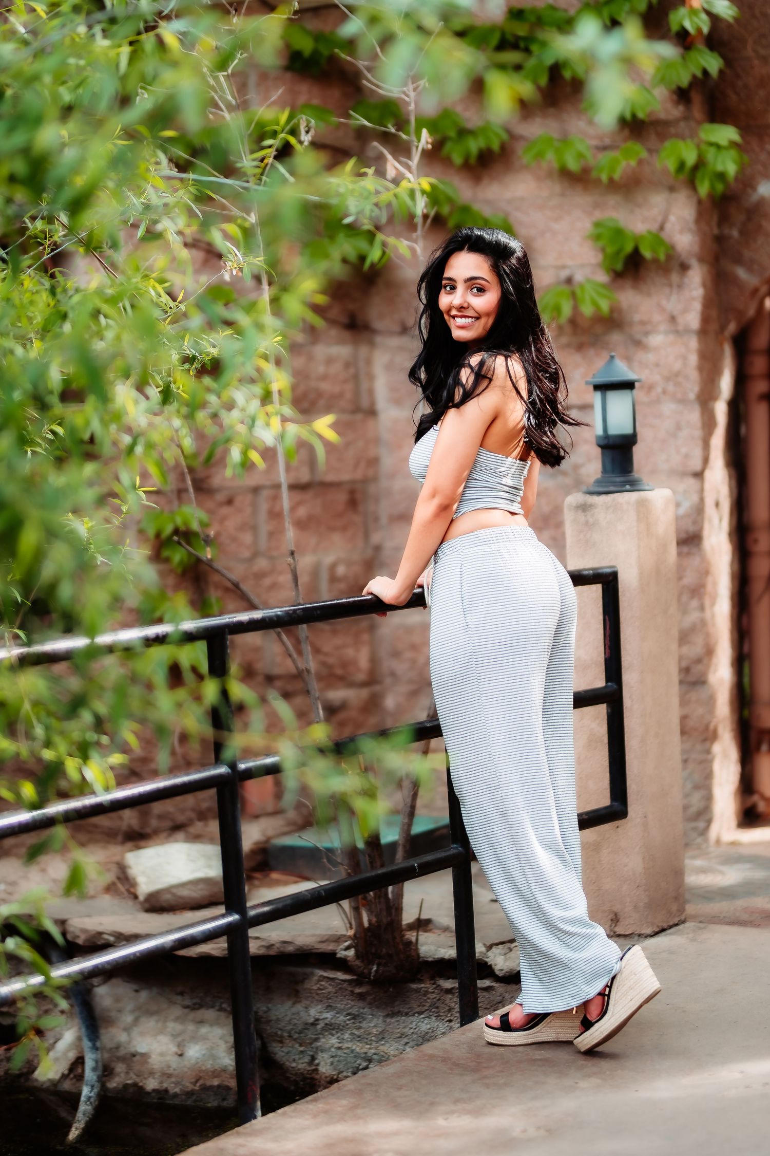 A person in a white jumpsuit poses gracefully on outdoor steps surrounded by green foliage.