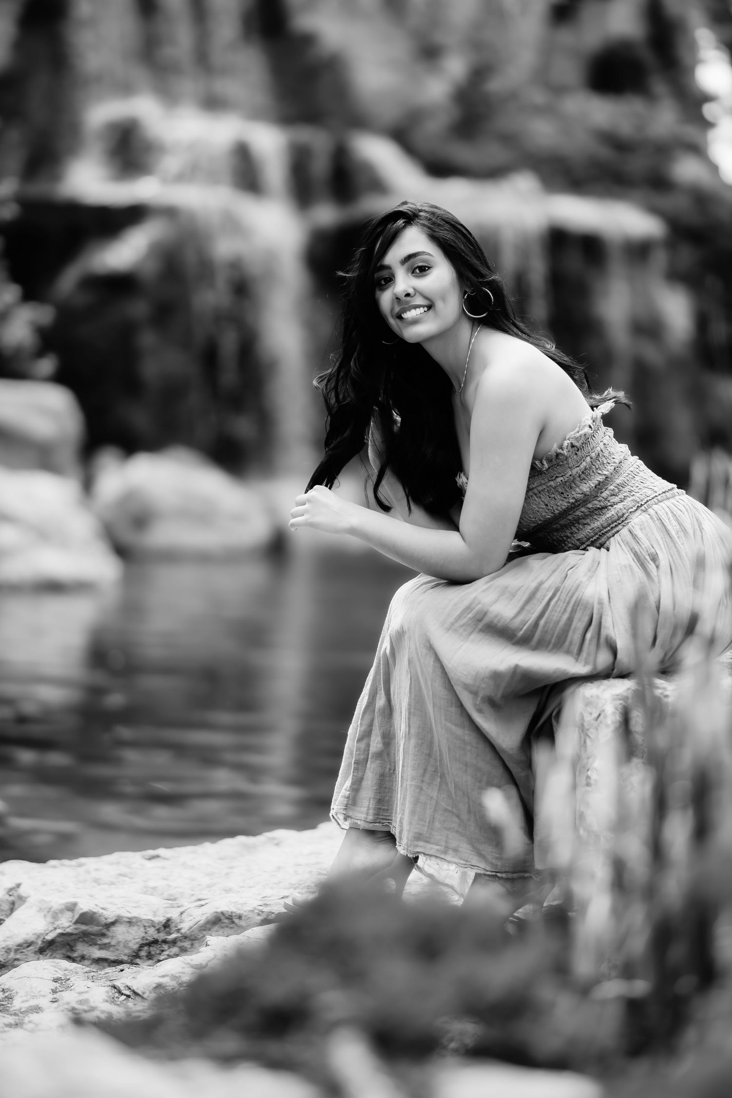 Black and white portrait of a person in a flowing dress sitting by a pond in a natural setting.
