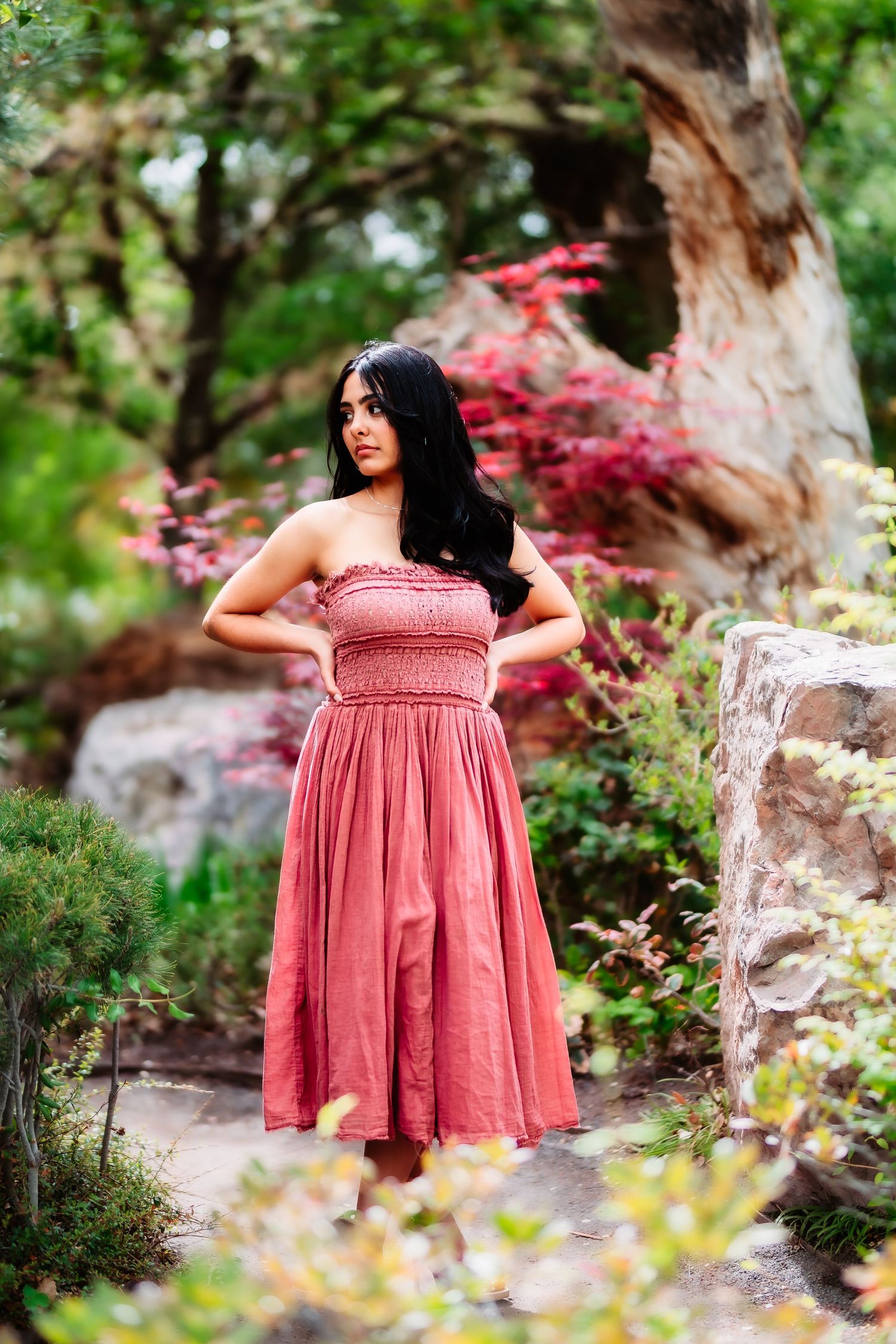 Woman wearing a pink sundress stands among red maple leaves and garden rocks.