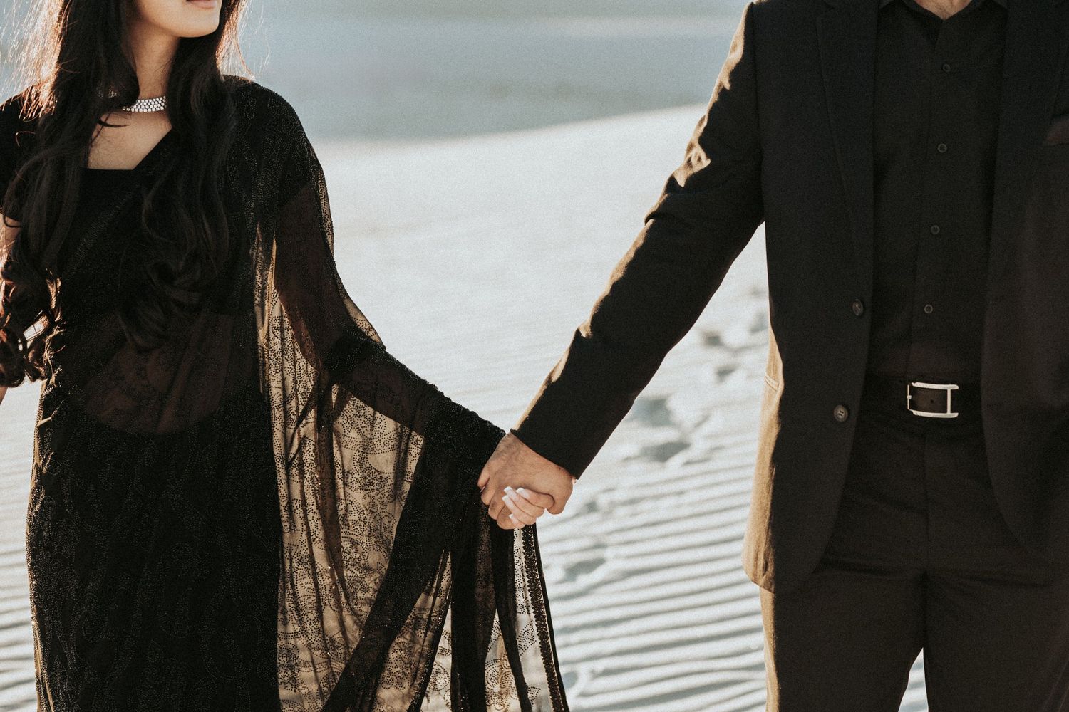 Couple in black attire holding hands while standing in white sand during a desert photoshoot.
