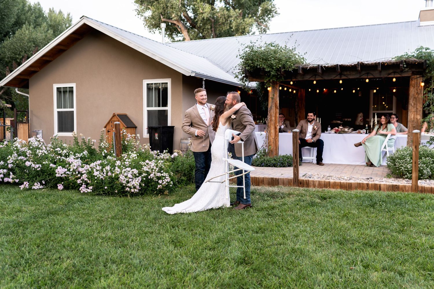 A wedding celebration outside a tan stucco house with string lights and floral decorations.