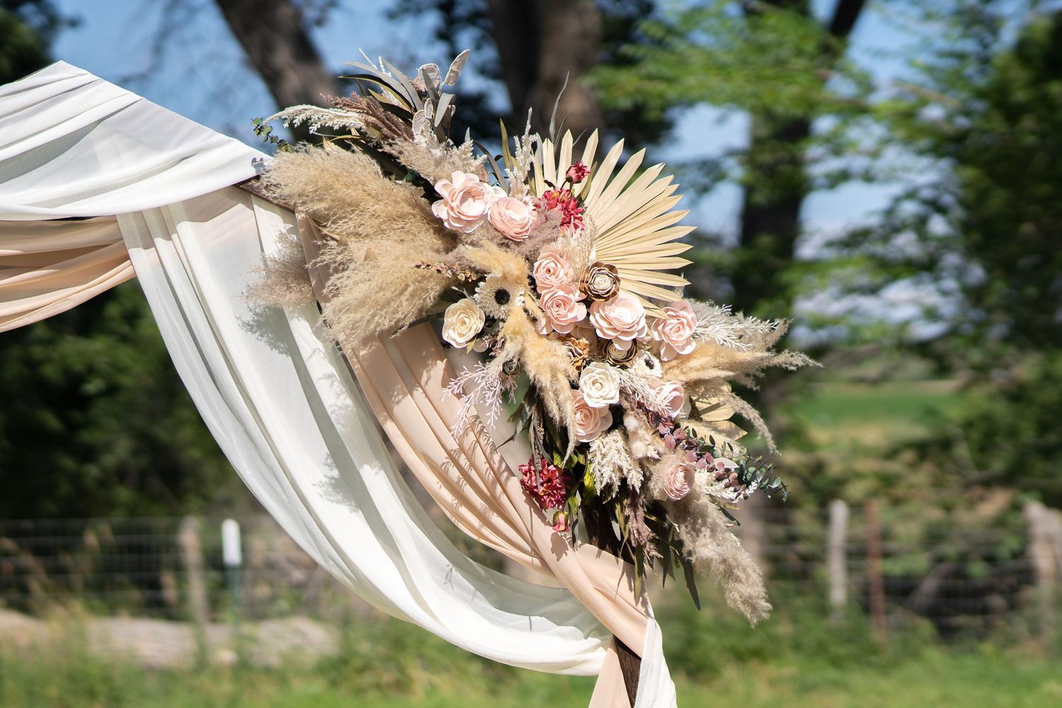 Rustic wedding arch corner decorated with dried pampas grass, blush pink flowers and white fabric in an outdoor setting.