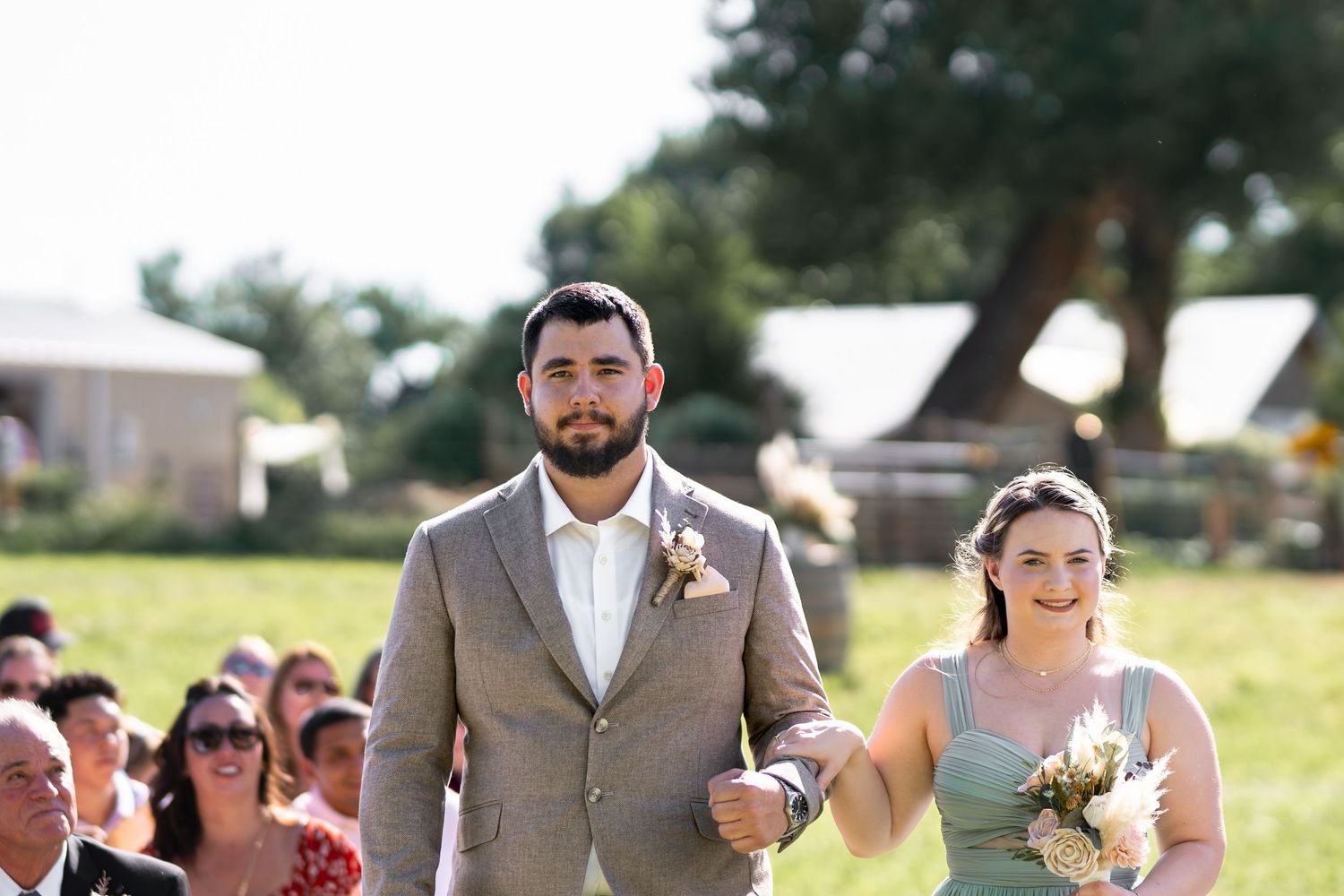 A couple proceeds down the aisle at an outdoor wedding with guests seated on white chairs.