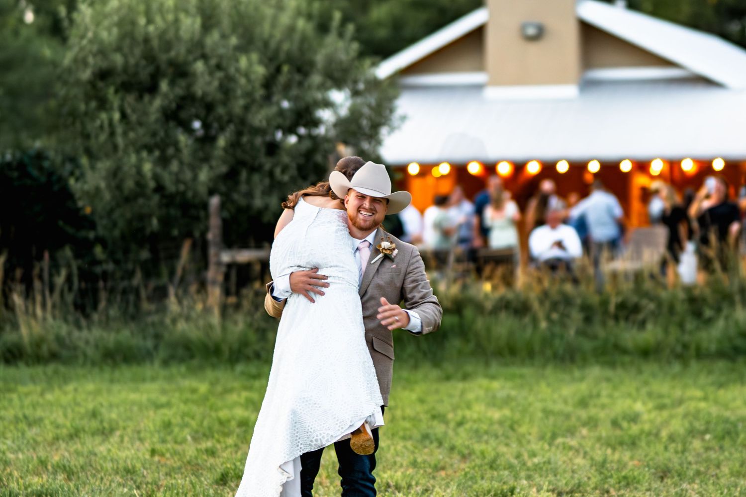 A couple embraces outdoors at a rural wedding with orange string lights and a rustic building in the background.