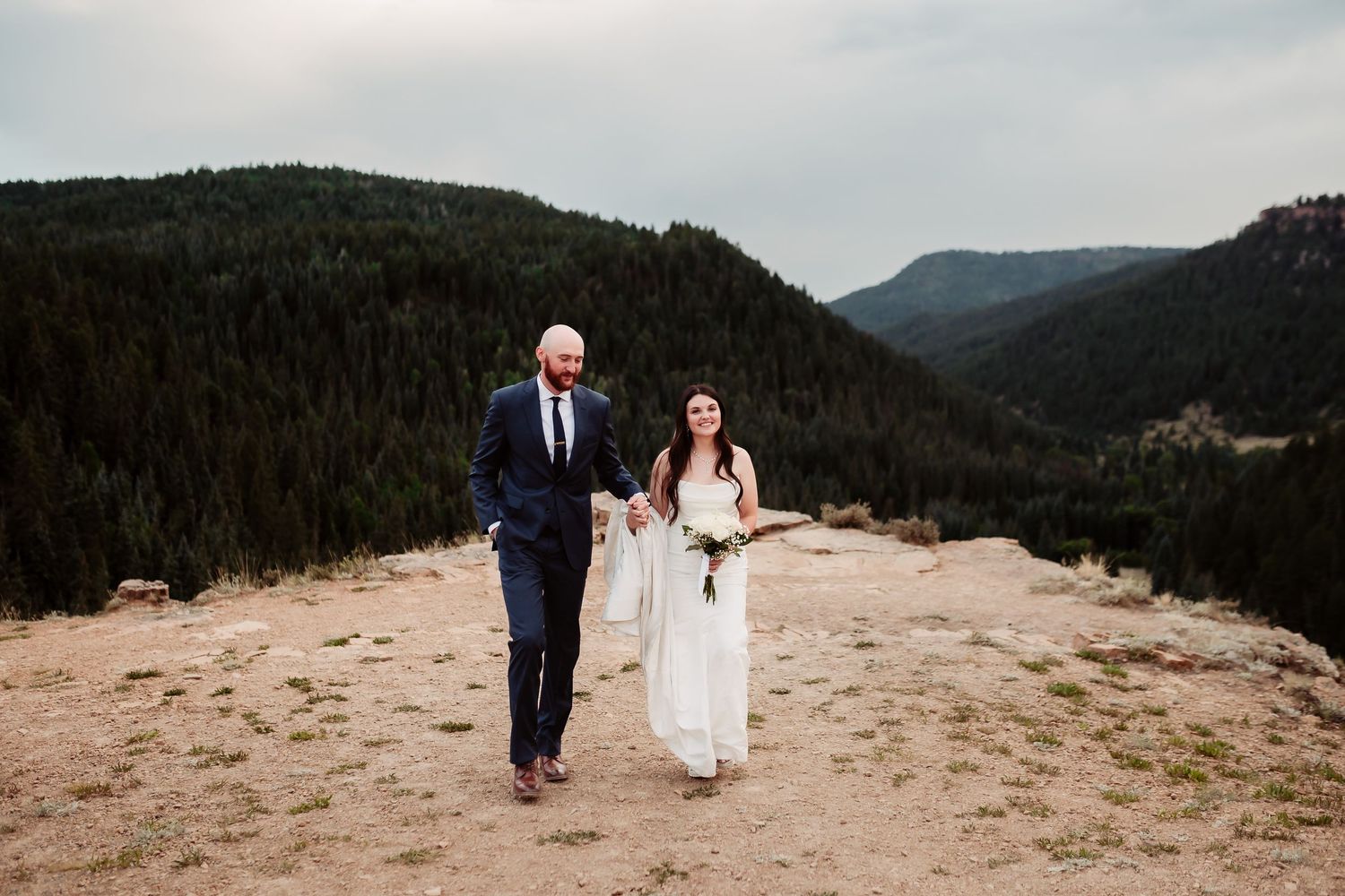 A newlywed couple walks together on a mountain trail with evergreen forests in the background.