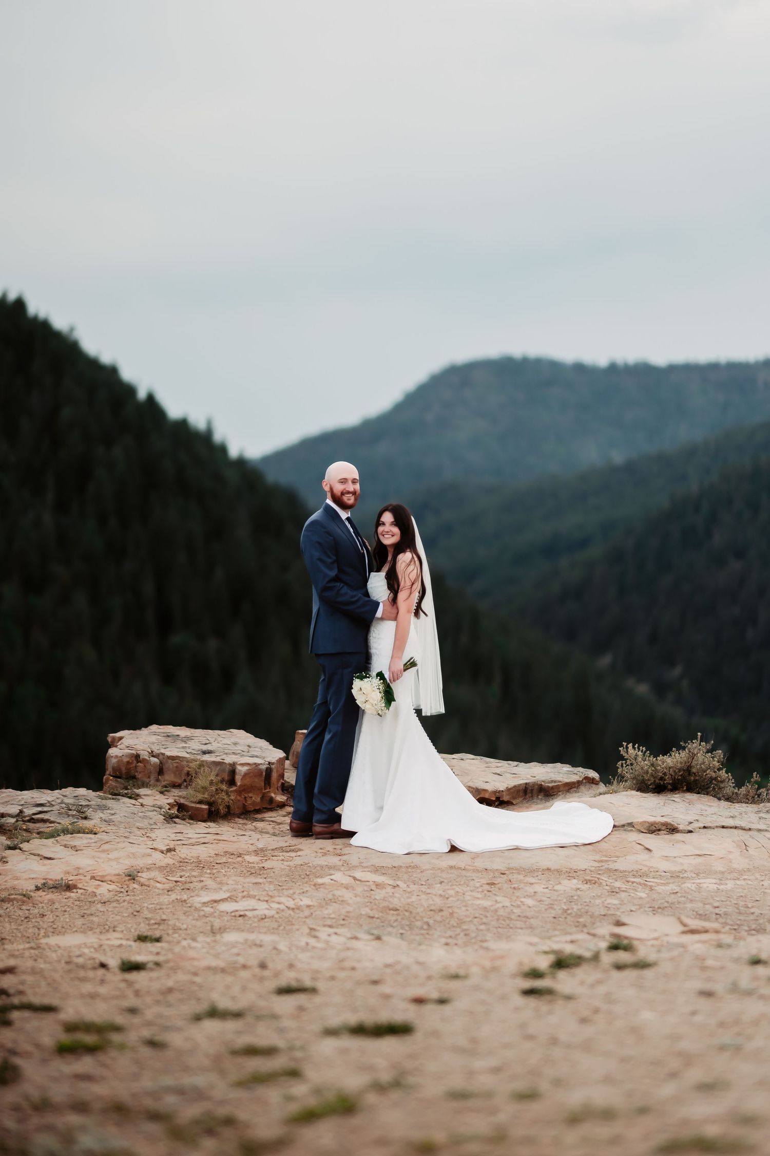 A couple in wedding attire poses on a rocky mountain ledge with sweeping forest views below.