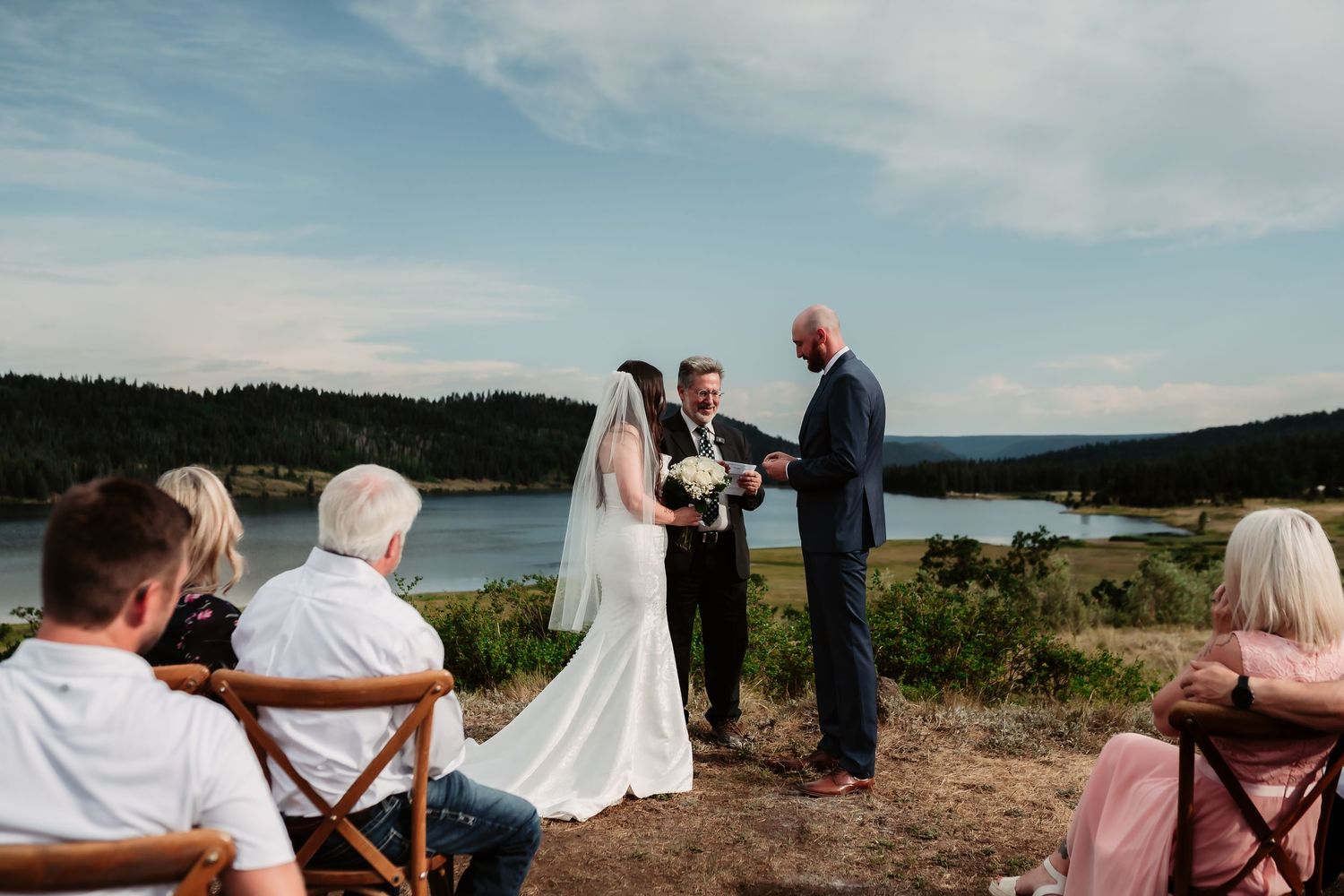 A scenic lakeside wedding ceremony with guests seated in wooden chairs on a natural overlook.