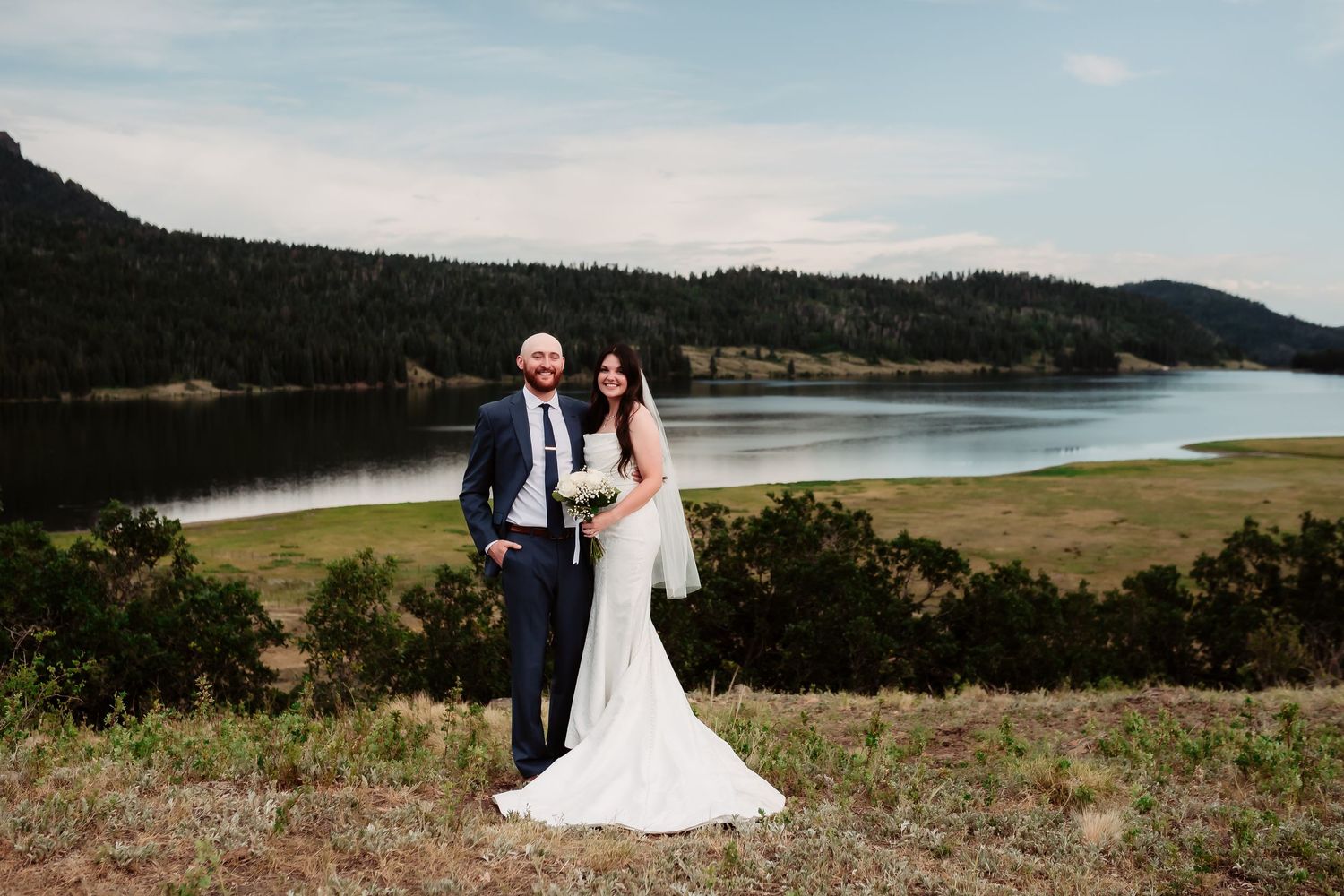 A newlywed couple poses for a portrait on a hillside with a scenic lake and mountains in the background.