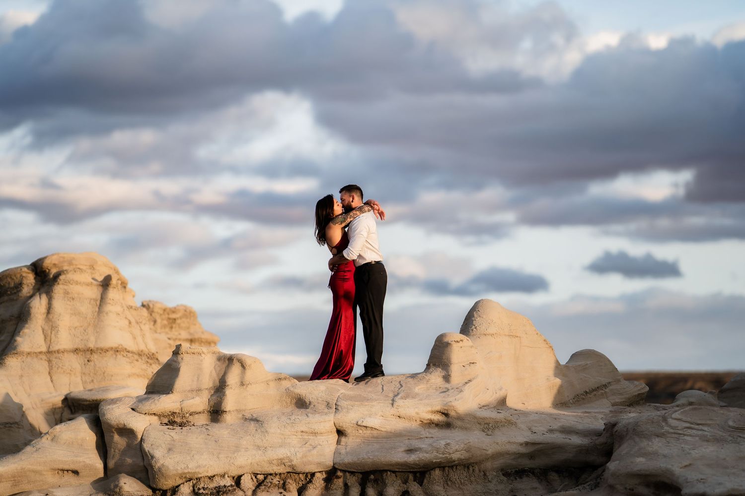 A couple embraces on rugged desert rock formations under dramatic cloudy skies.