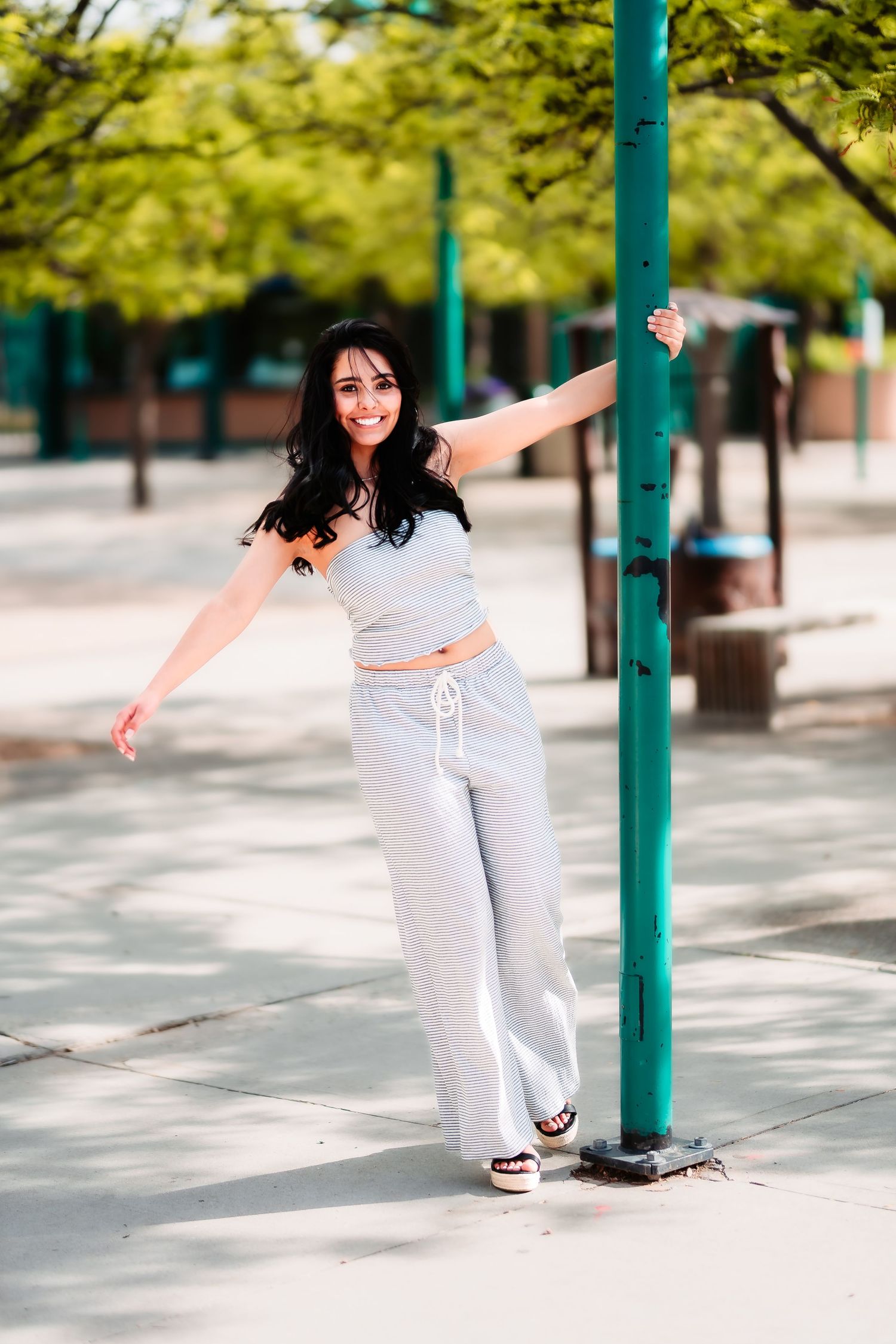 A person in casual white attire poses playfully while holding onto a green pole in an urban setting.
