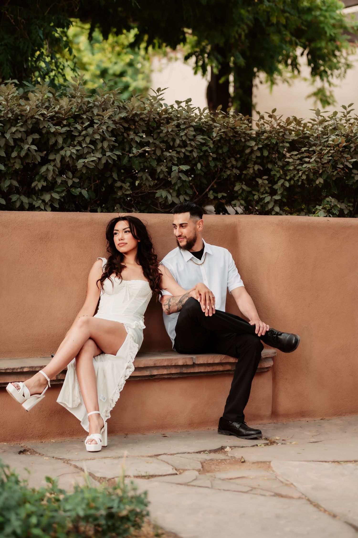 A stylish couple sits together on a terracotta-colored wall, sharing playful and intimate moments.