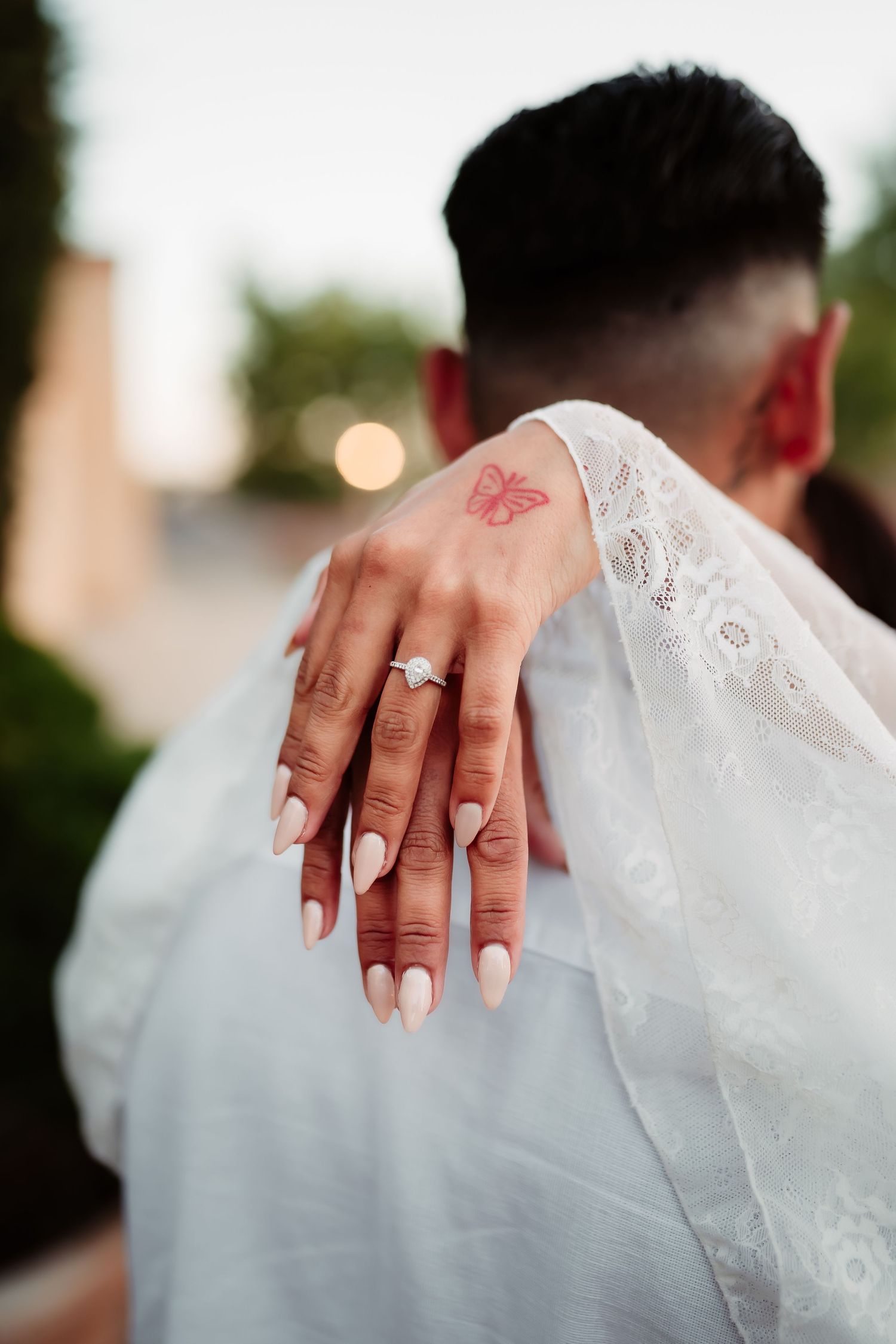 A close-up of a hand with white nail polish and an engagement ring against lace fabric with a butterfly tattoo.