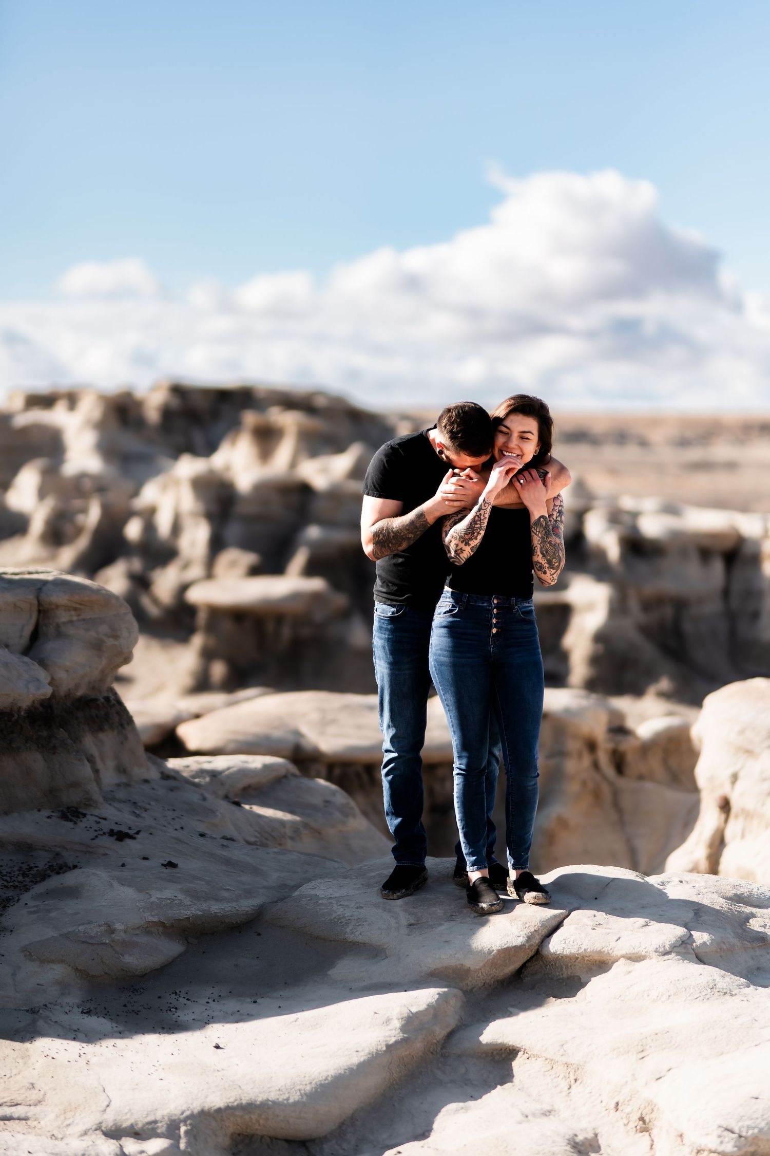 Two people embrace while standing on rocky white terrain against a blue sky with puffy clouds.