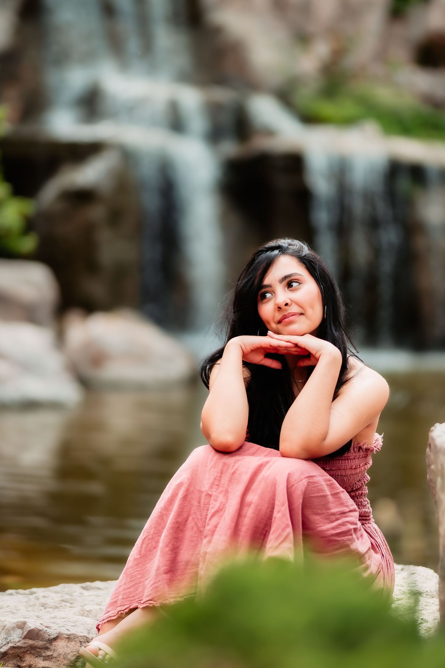 Person in a coral dress sits contemplatively by a scenic waterfall on rocks in a peaceful nature setting.
