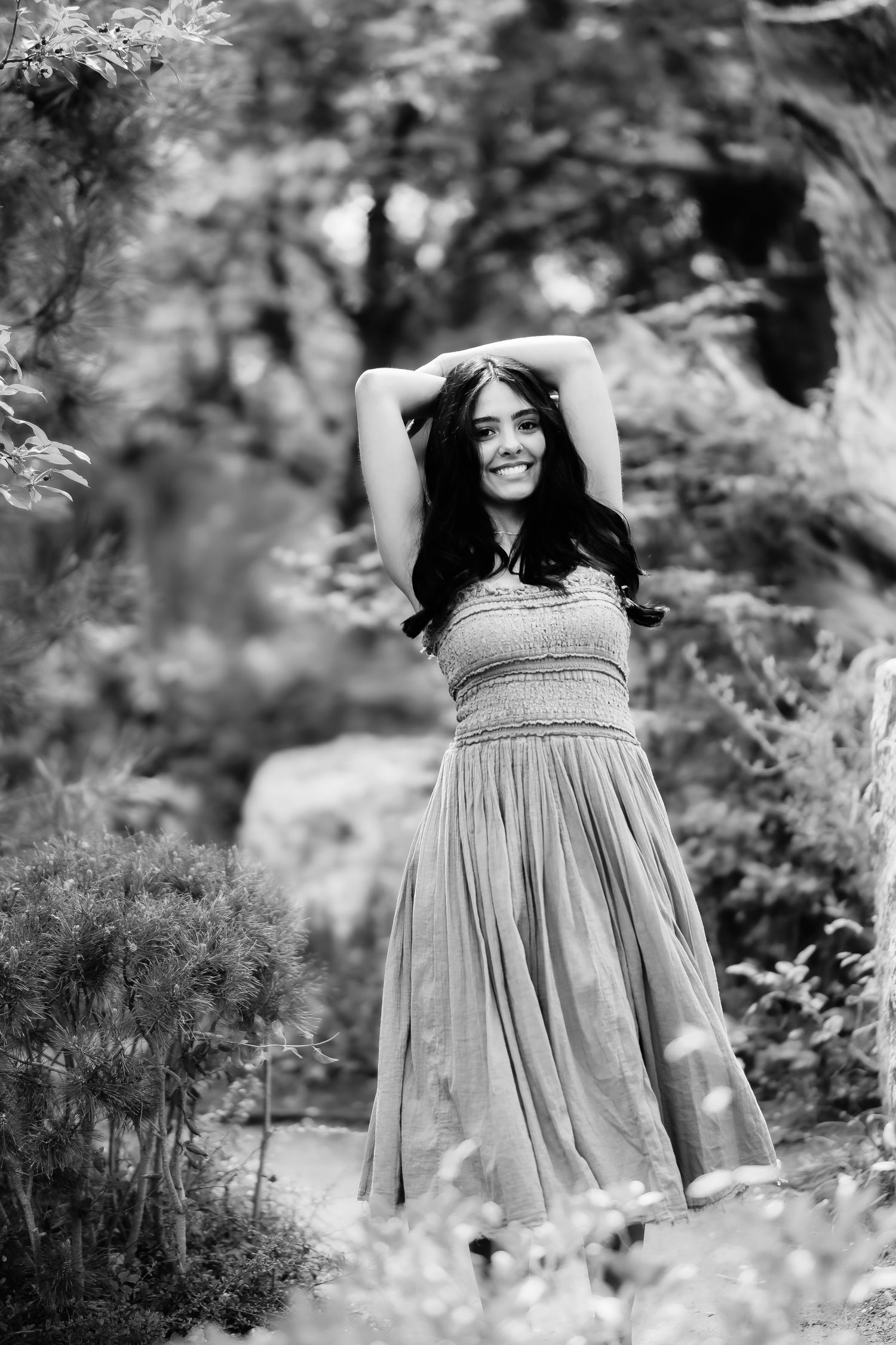 Woman in flowing dress poses with raised arms amidst garden foliage in black and white photograph.