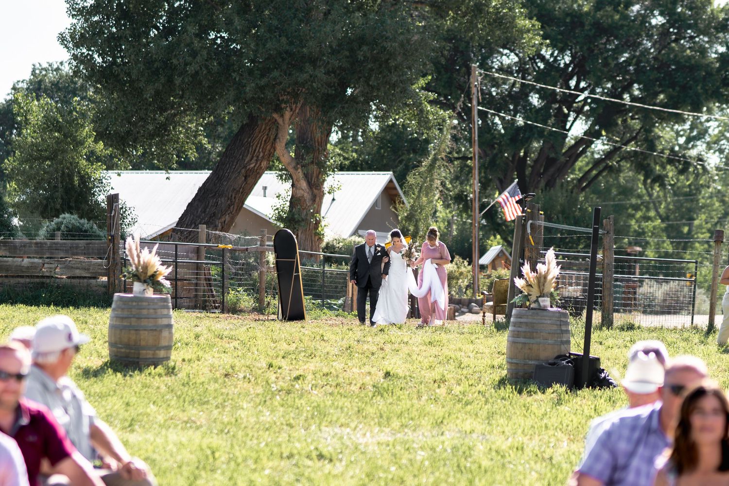 Wedding guests gather under trees at an outdoor wedding ceremony on a sunny day.