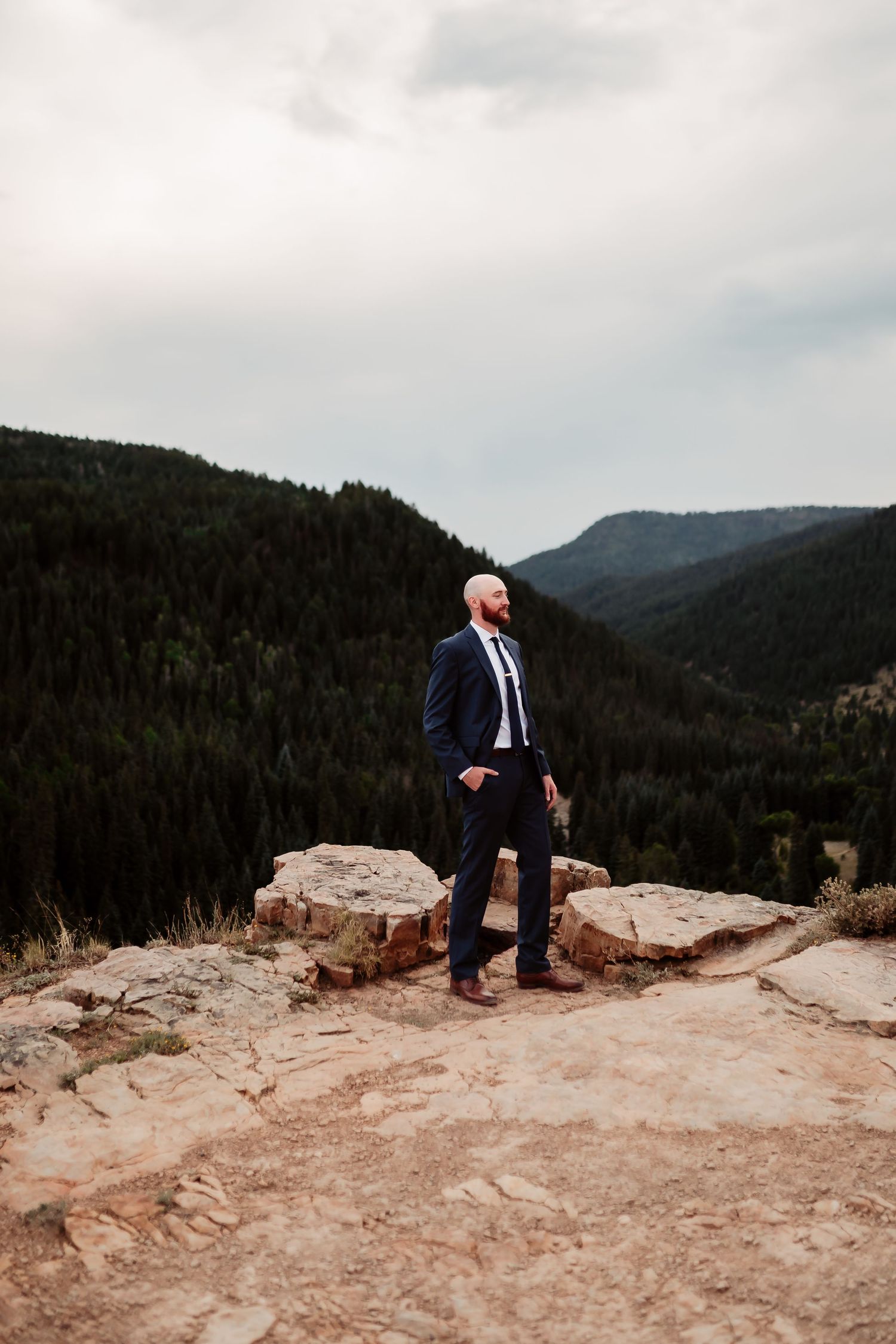 Groom exploring rocky mountain terrain while wearing a navy suit with scenic mountain vistas in background.