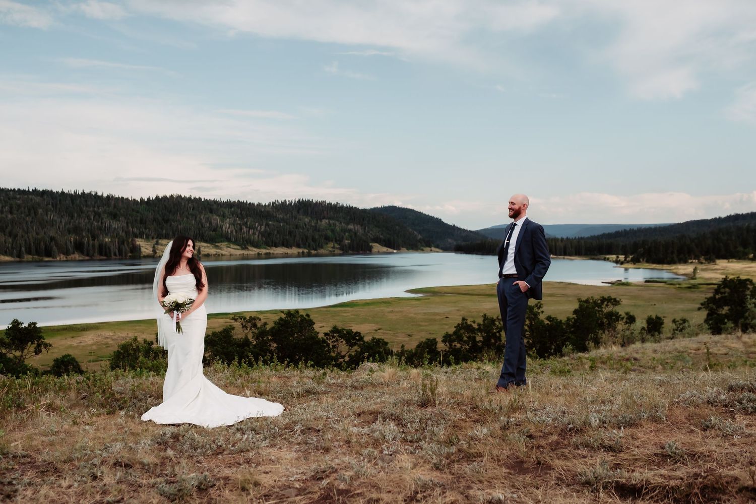 A wedding couple poses separately on a hilltop with a stunning lake view in the background.