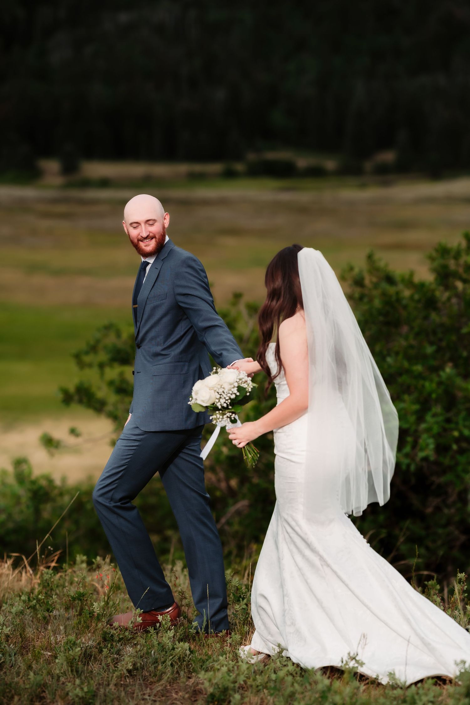 A playful wedding couple poses on a hillside with the bride holding a white bouquet.