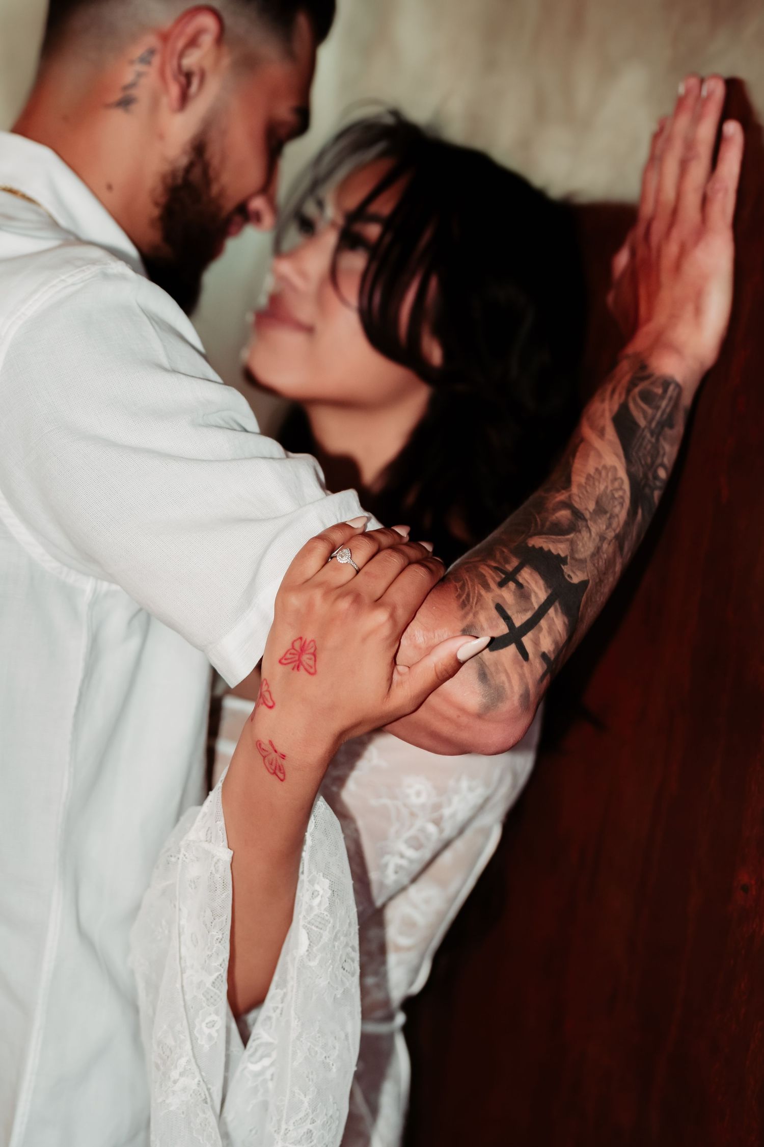 An intimate moment captured showing tattooed arms and formal white attire against a wall.