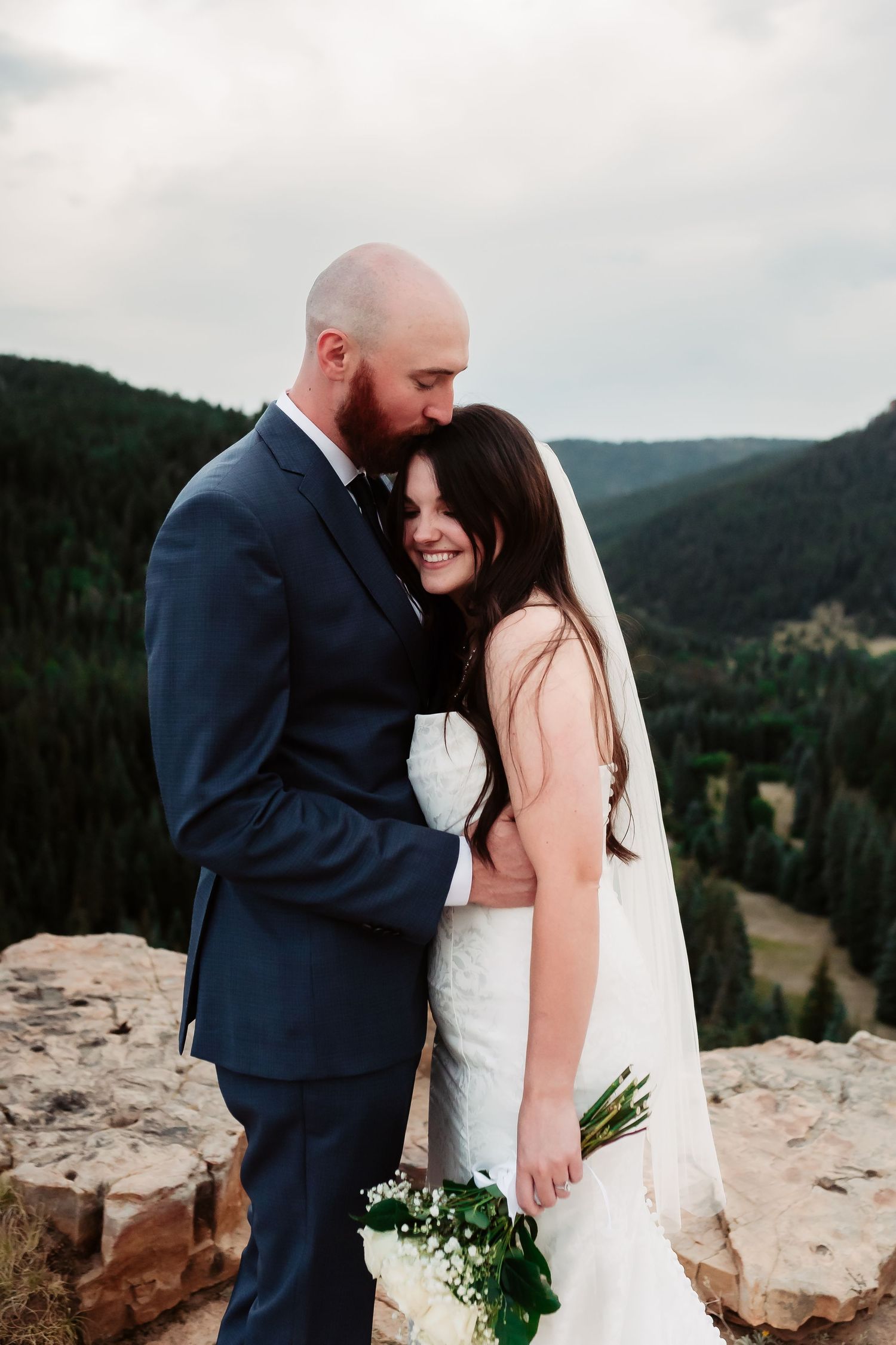 Tender embrace between newlyweds on rocky mountain ledge with scenic valley views.