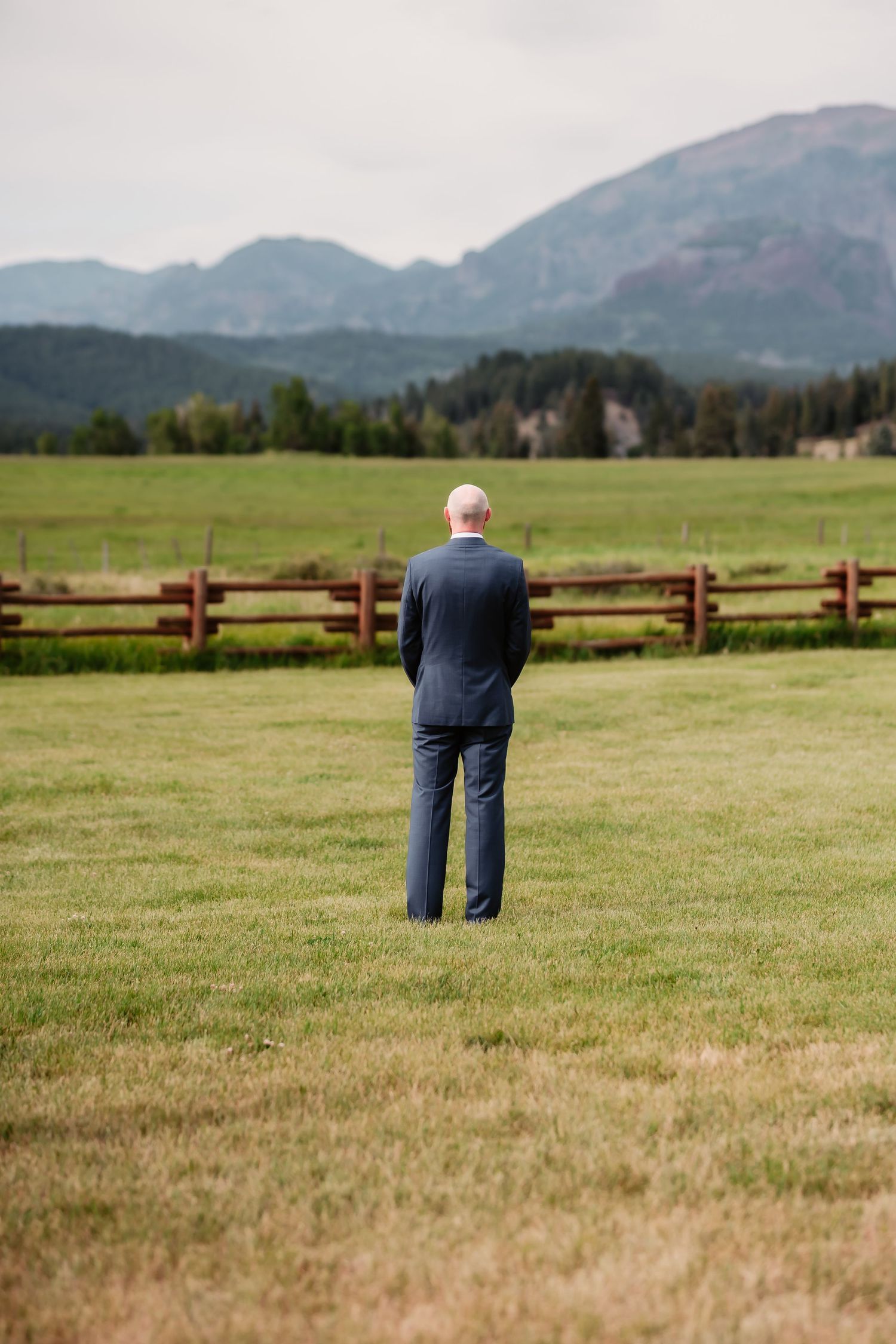 Person in casual wear stands alone in a mountain meadow with wooden fence and rolling hills in view.
