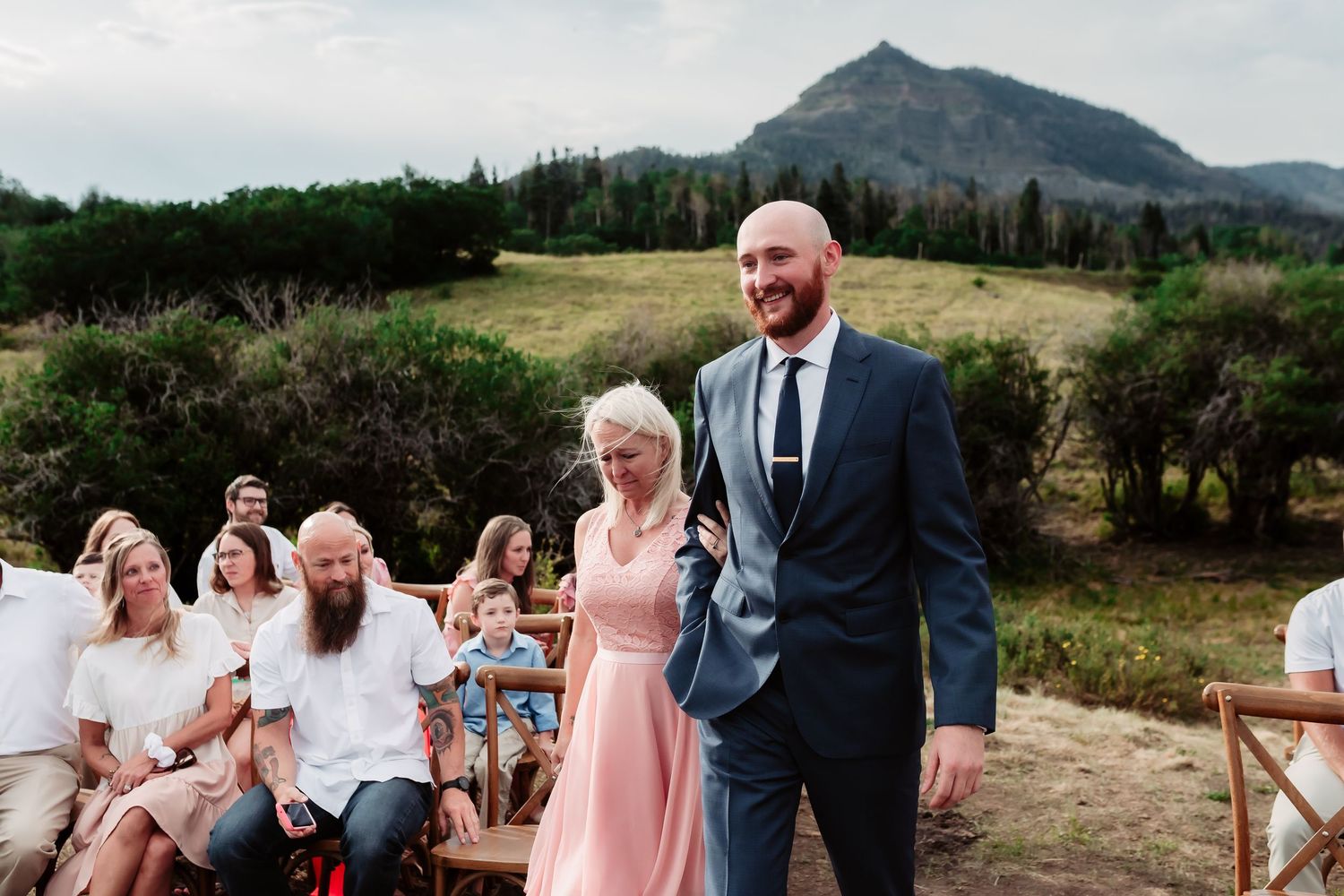 Wedding ceremony guests seated on rustic chairs with mountain peaks in background.