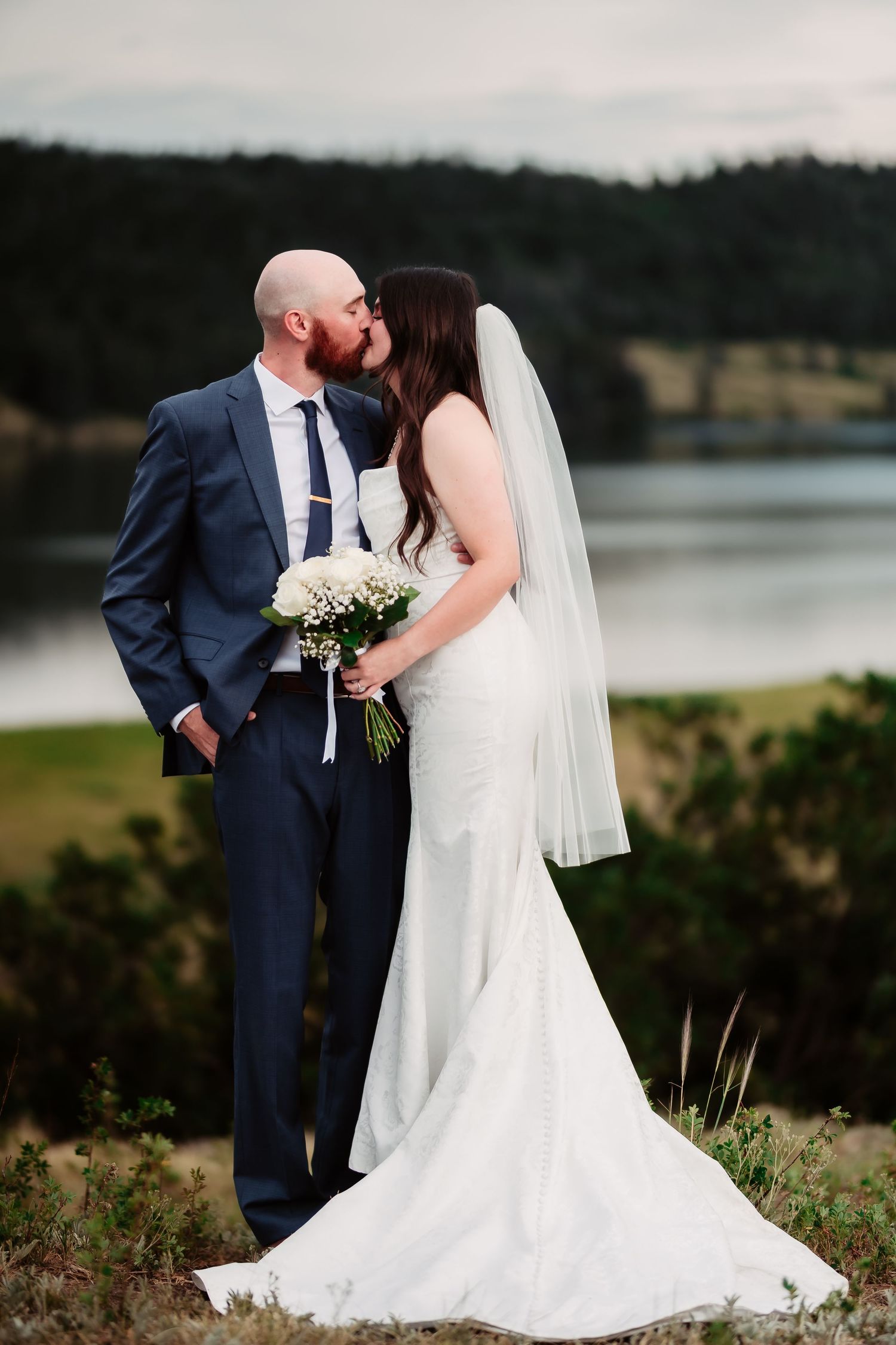 Wedding couple poses near lake with bride in flowing white dress and groom in navy suit.