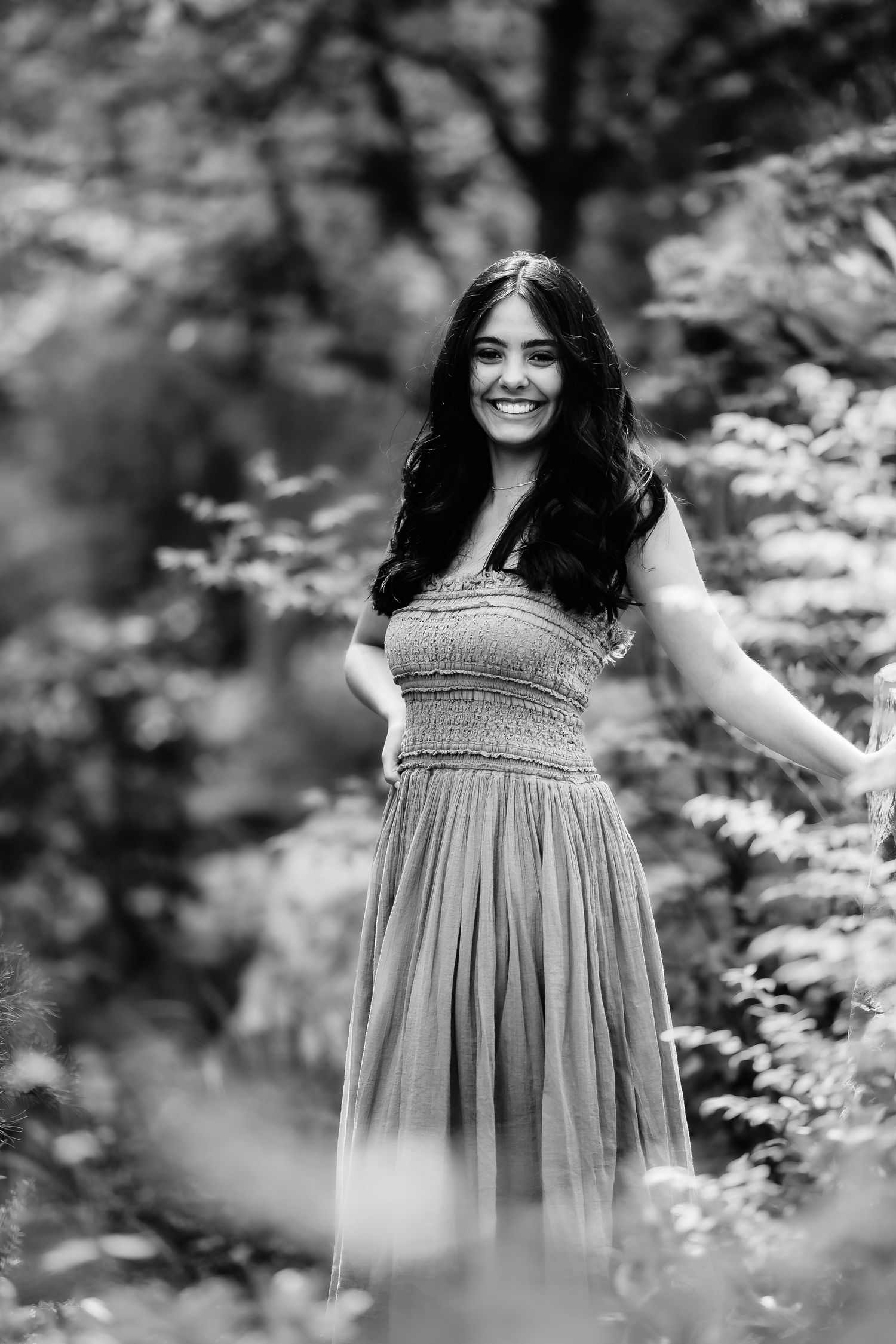 A black and white portrait of a person in a flowing dress smiling outdoors in a natural setting.
