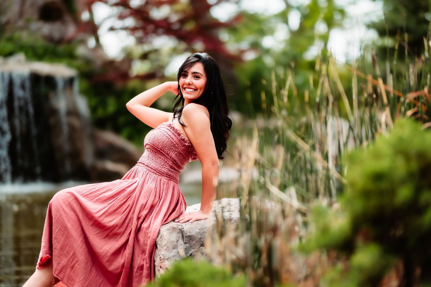 A person in a flowing coral dress poses gracefully by a garden waterfall in an outdoor photoshoot setting.