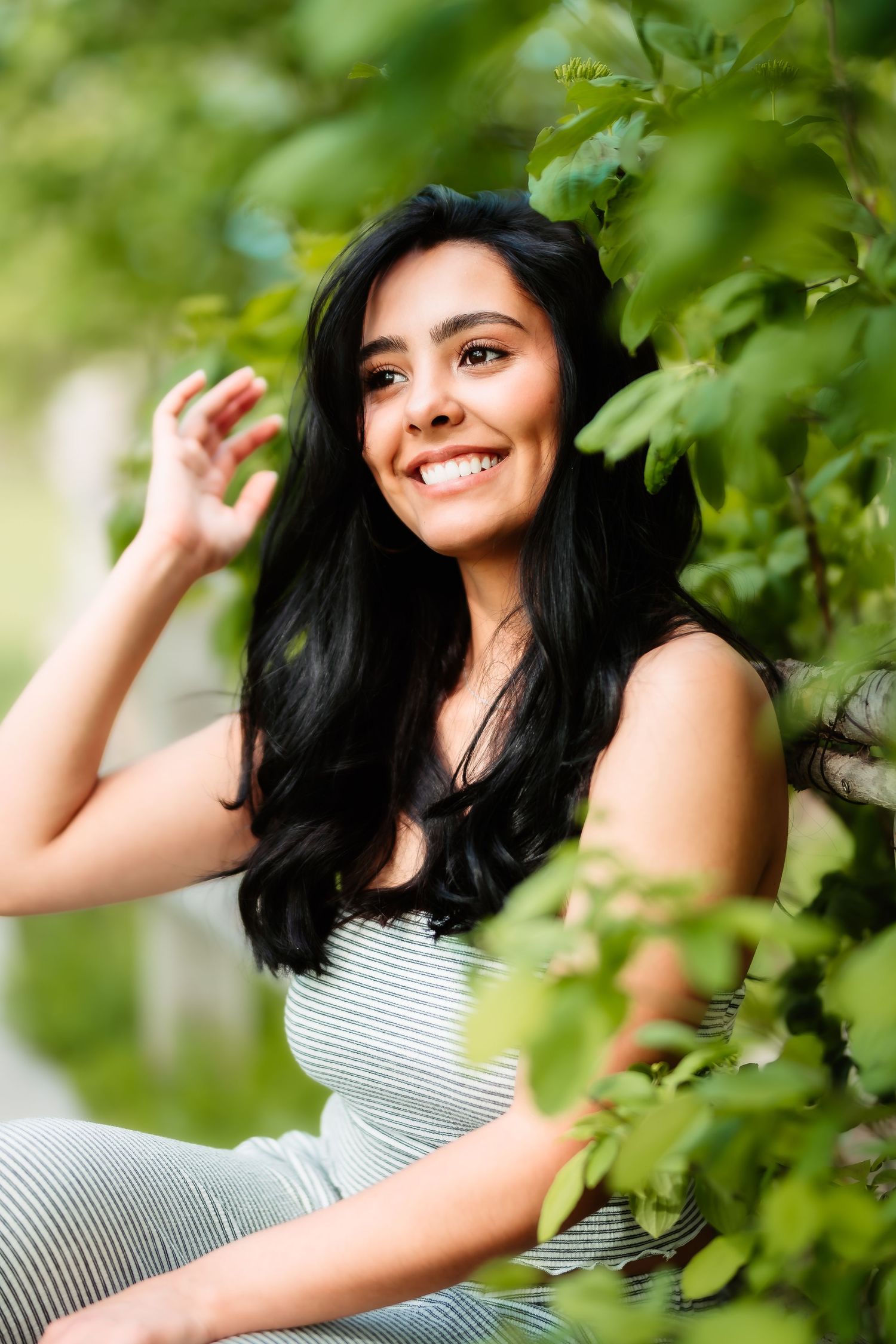 Portrait of long-haired woman in striped top smiling joyfully among green leaves in natural outdoor setting.