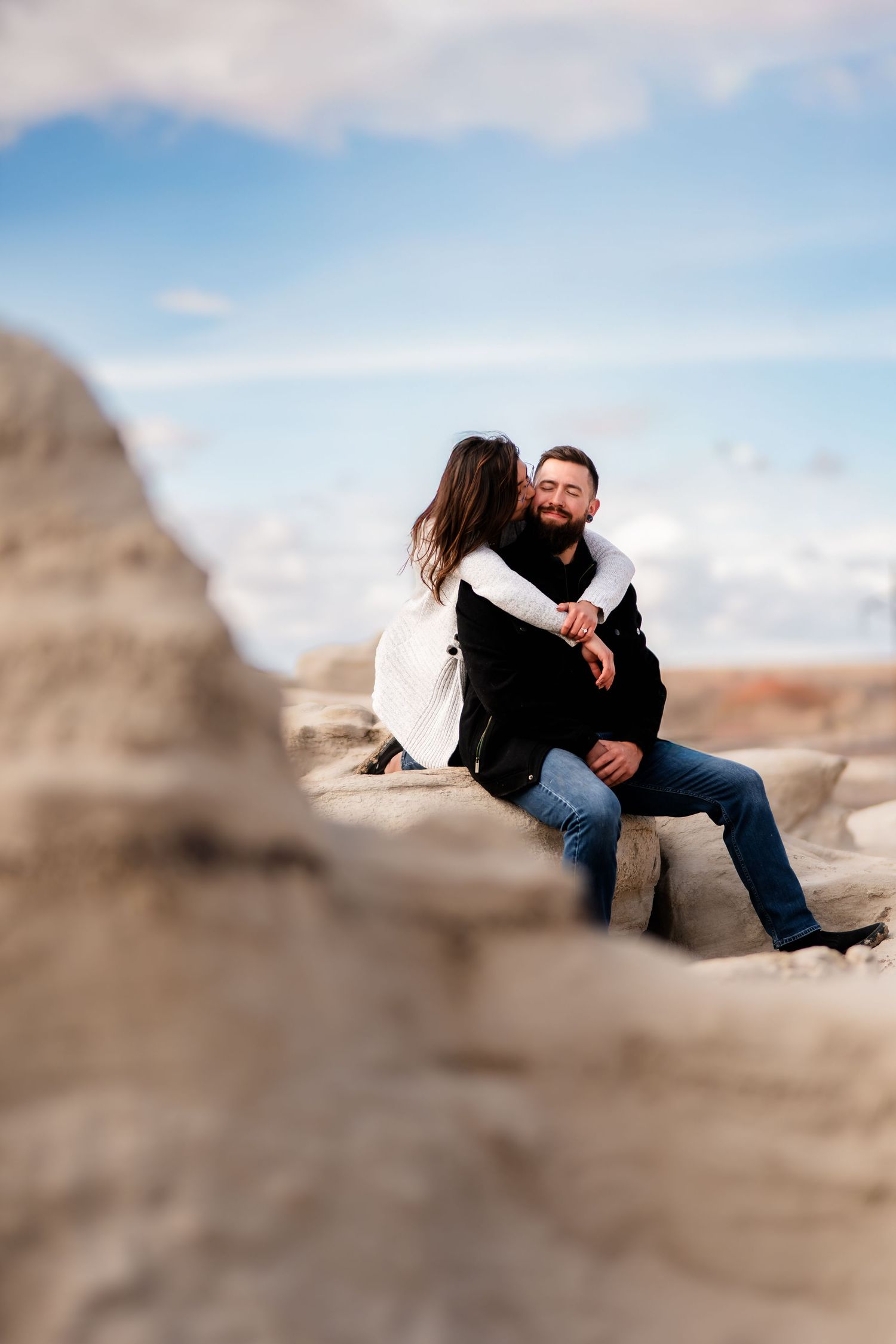 A couple shares an intimate moment while sitting on rocky terrain beneath a blue sky.