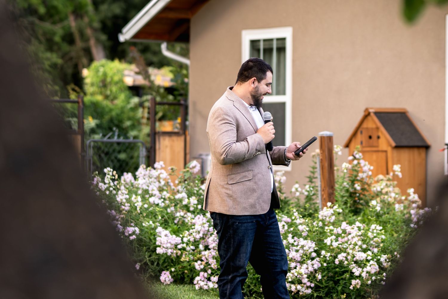 A person in a beige blazer stands among blooming white flowers while looking at their phone in a garden setting.