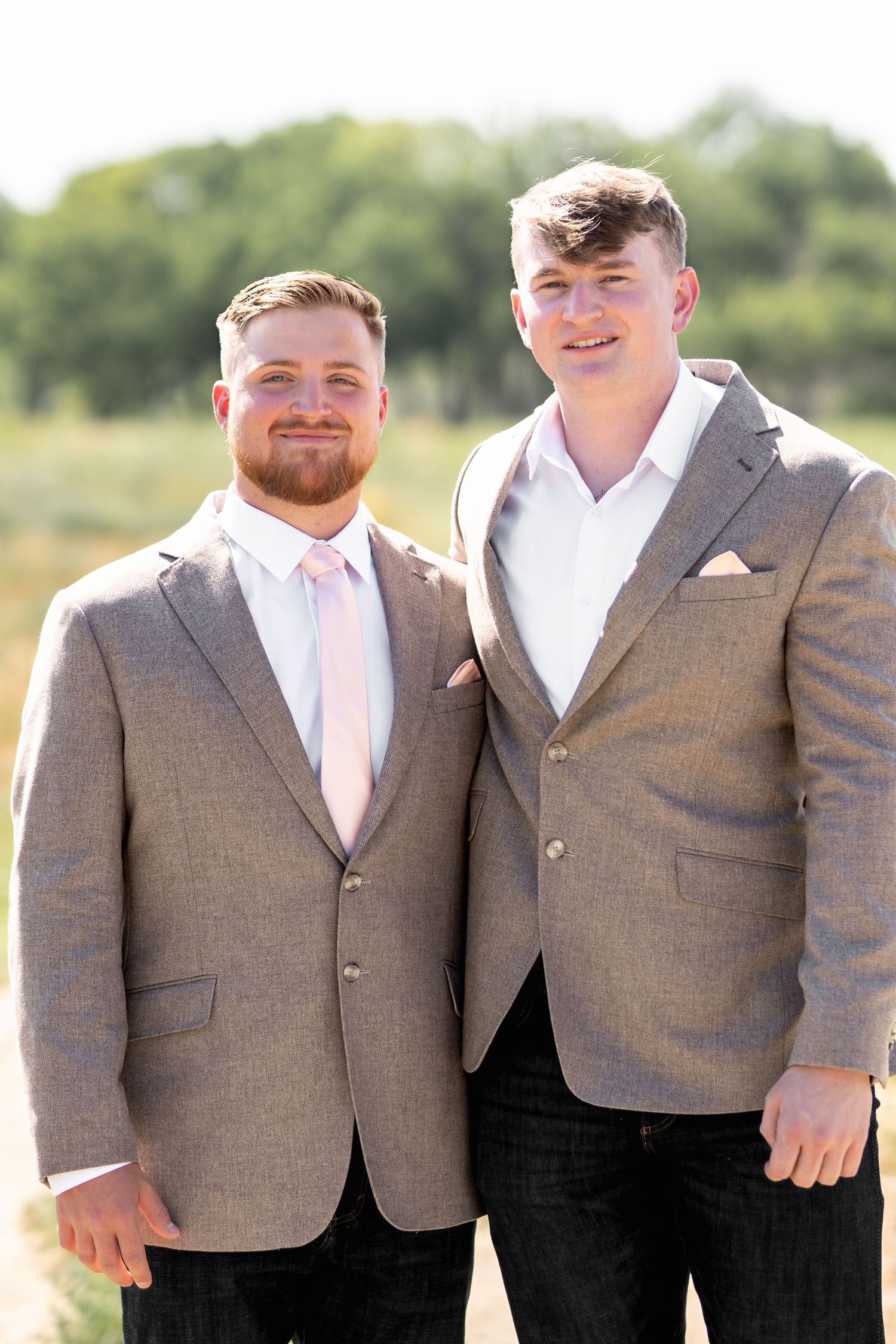 Two groomsmen in matching gray suit jackets and white shirts pose together outdoors at a wedding.