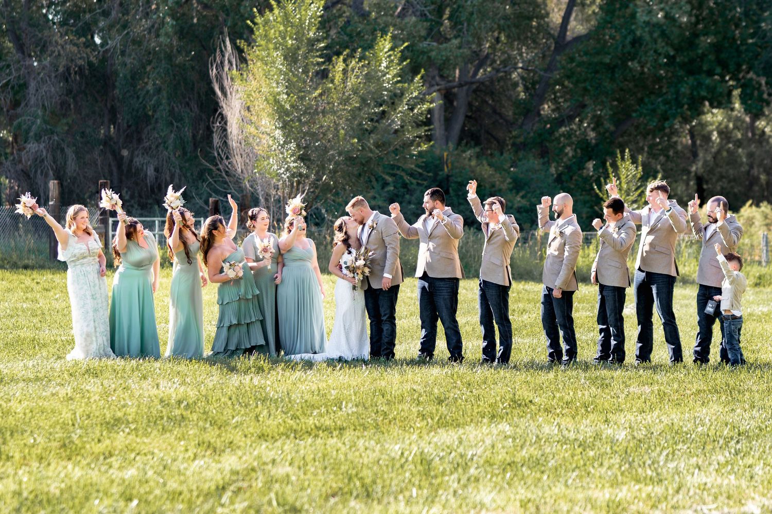 Wedding party in turquoise dresses and tan suits lined up for a fun group photo in a grassy field.