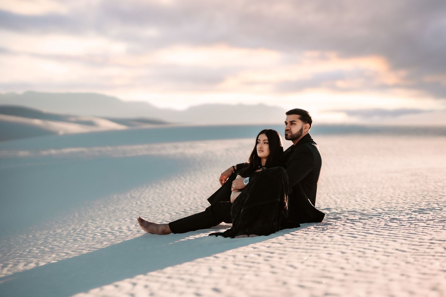 A couple sits together on pristine white sand dunes against a cloudy sunset sky.