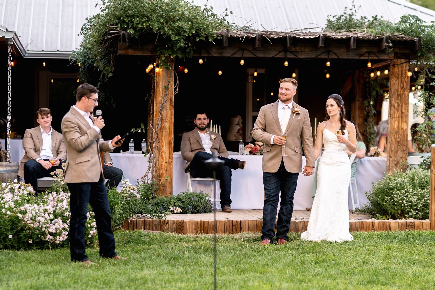 Outdoor wedding ceremony takes place under rustic wooden pergola with floral arrangements and string lights on a grassy lawn.