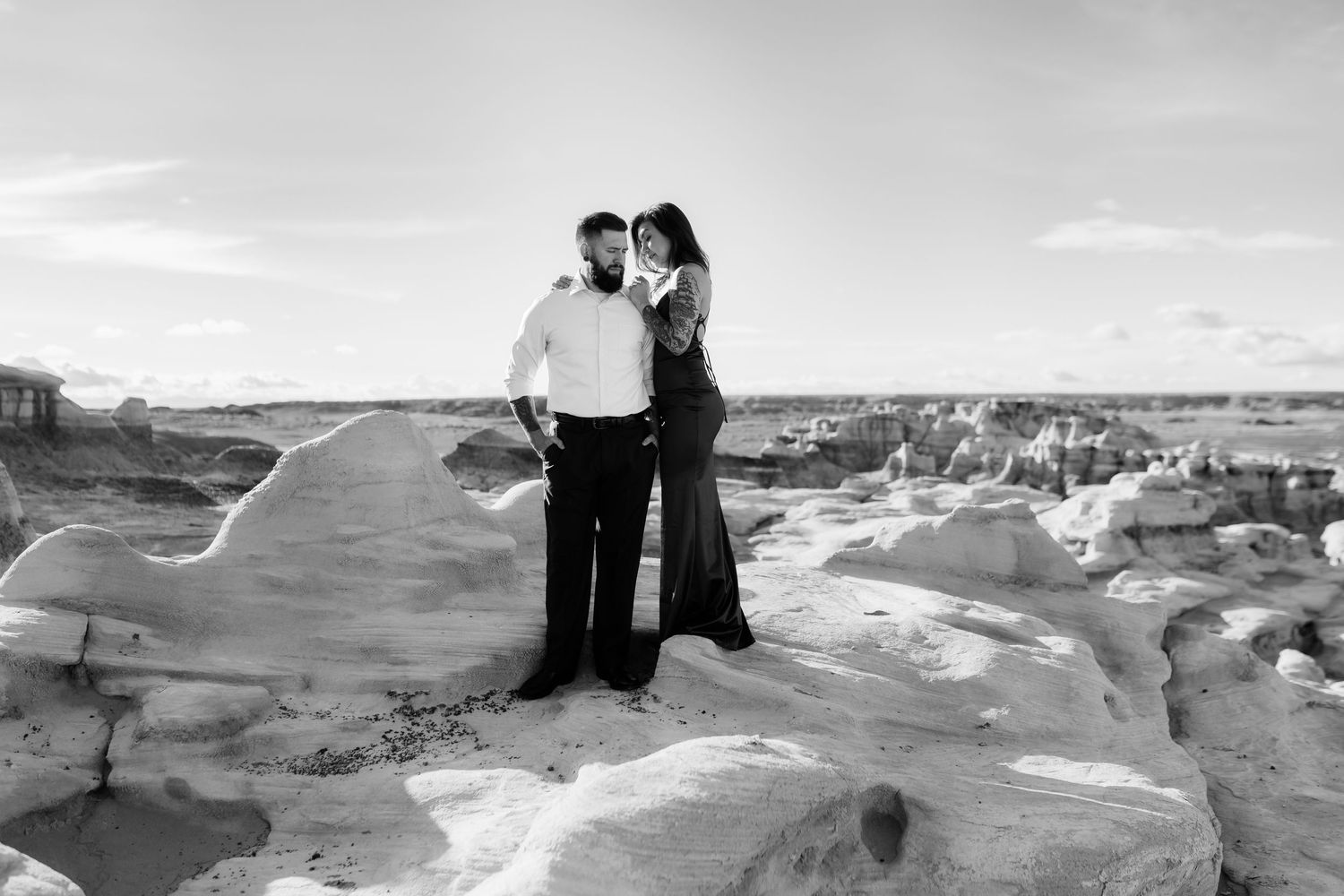Couple embraces on rugged rock formations in black and white desert landscape.