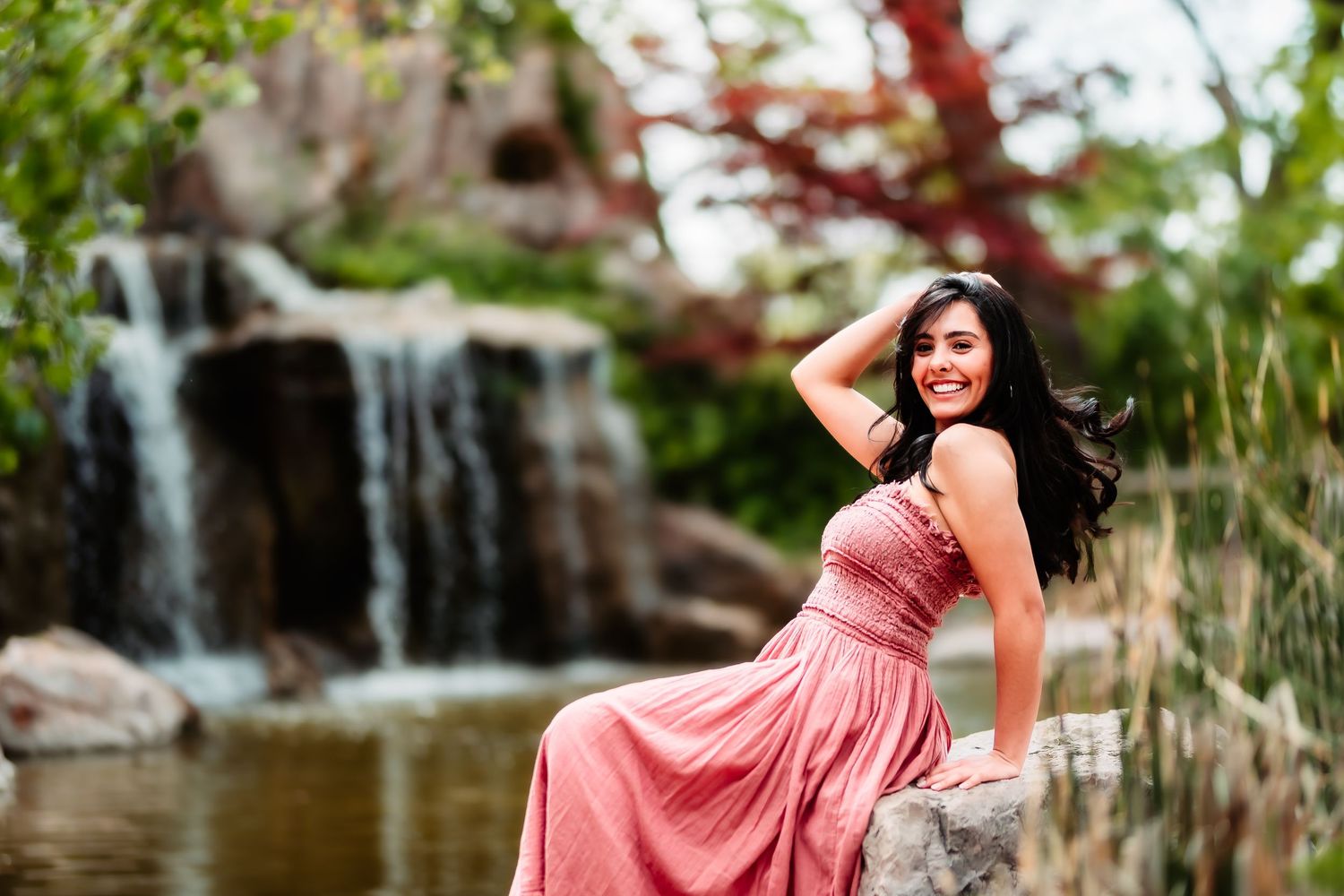 A woman in a rose-colored dress smiles while posing against a waterfall backdrop.