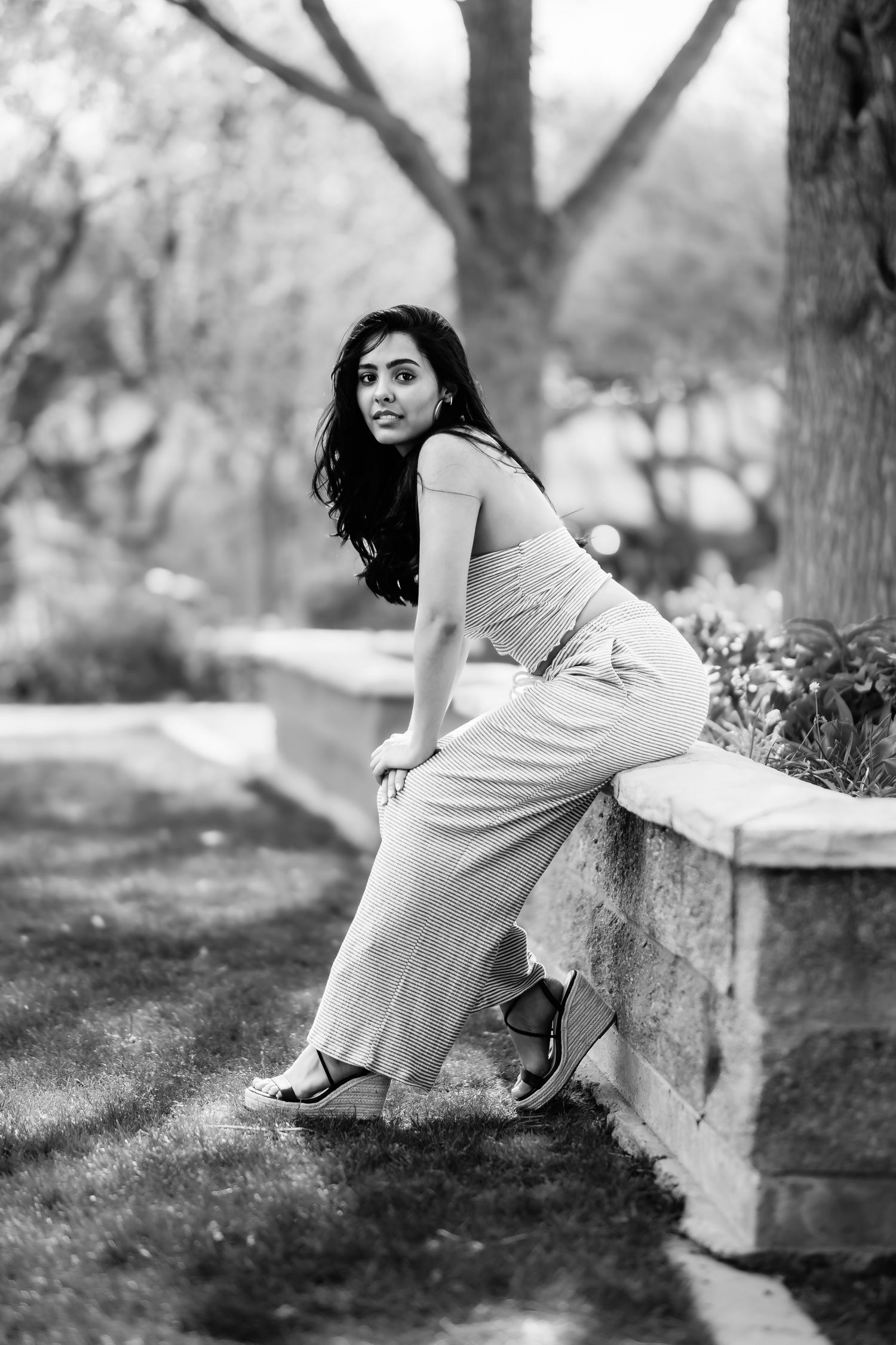 Black and white photo of woman in elegant dress leaning on stone wall in park setting.