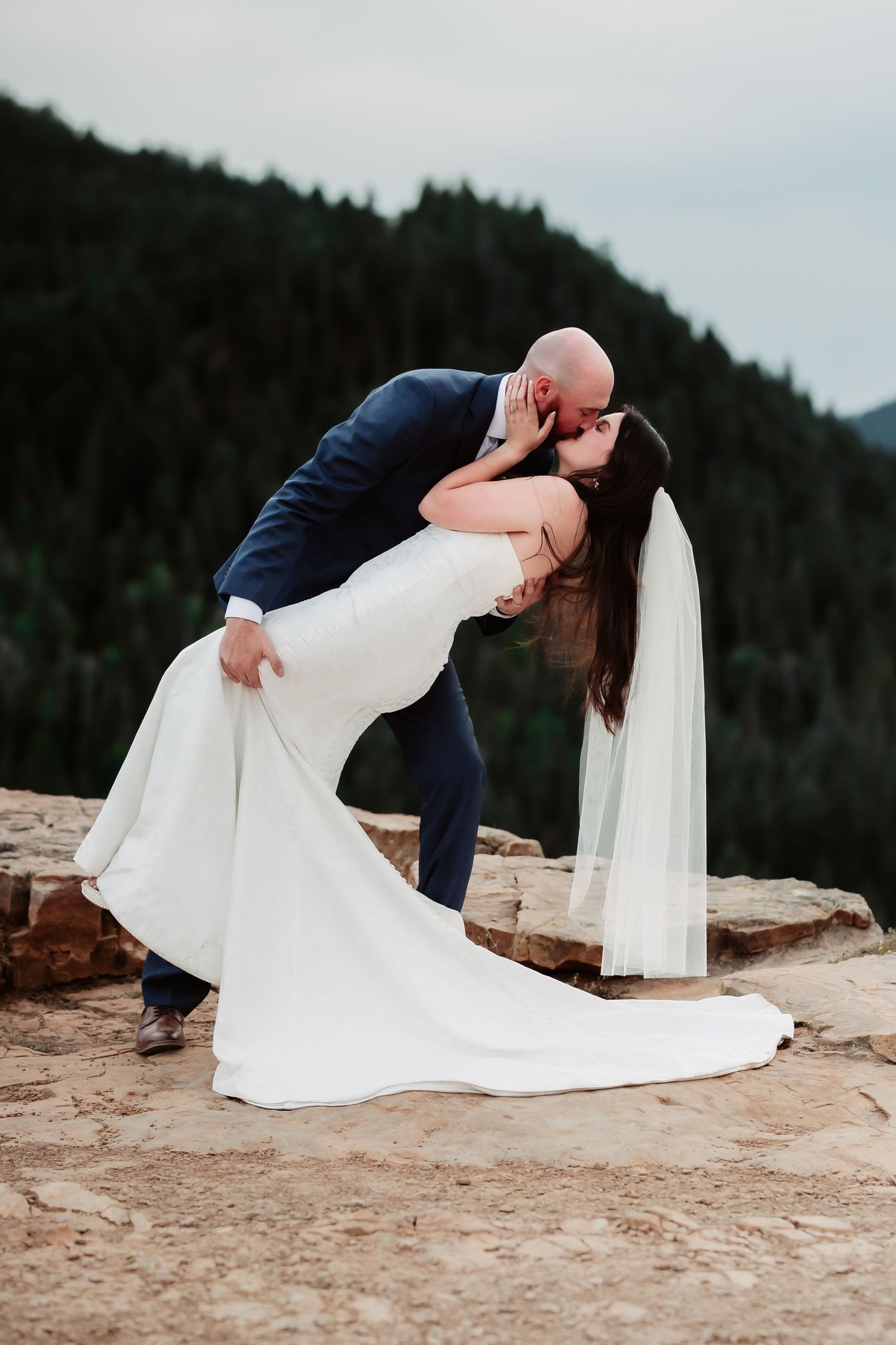 Romantic dip pose captured on rocky mountain ledge during sunset wedding photoshoot.