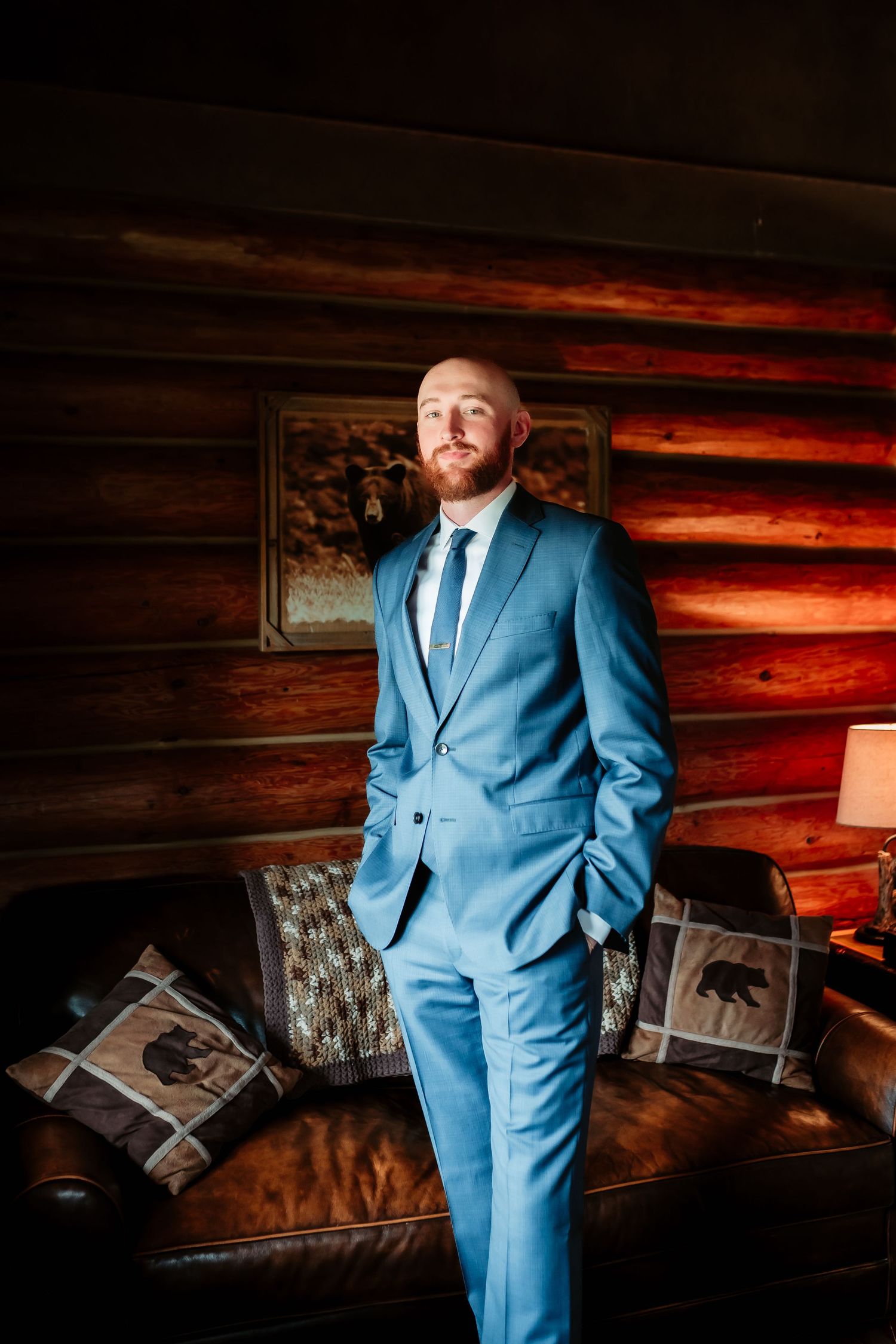 A person wearing a blue suit stands against rustic wooden cabin walls in dramatic lighting.