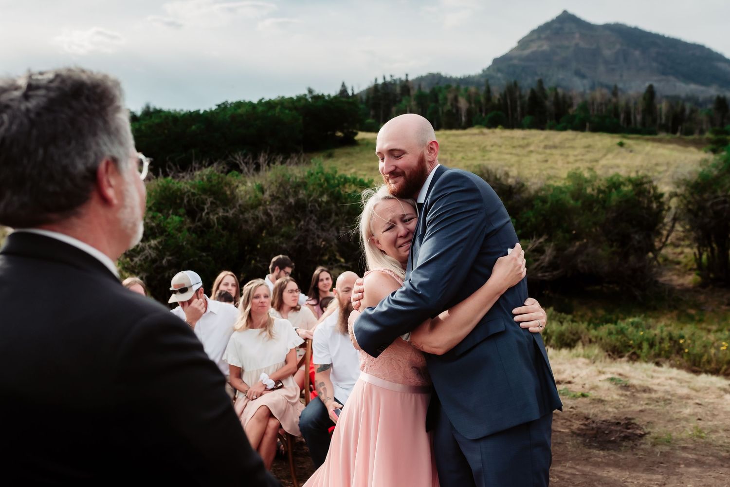 Wedding guests celebrate as couple embraces during outdoor ceremony with mountain backdrop.